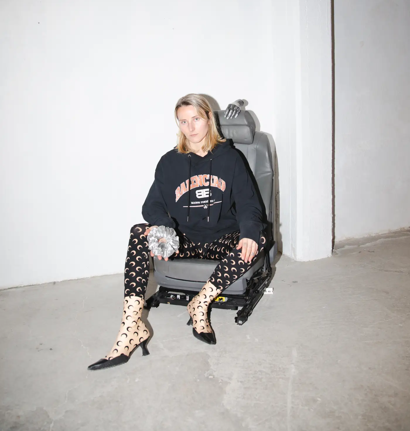Bety Krňanská sitting on a detached car seat sculpture in a white-walled gallery space, wearing crescent moon tights and heels, holding a metal object with a prosthetic hand resting on the seat behind her