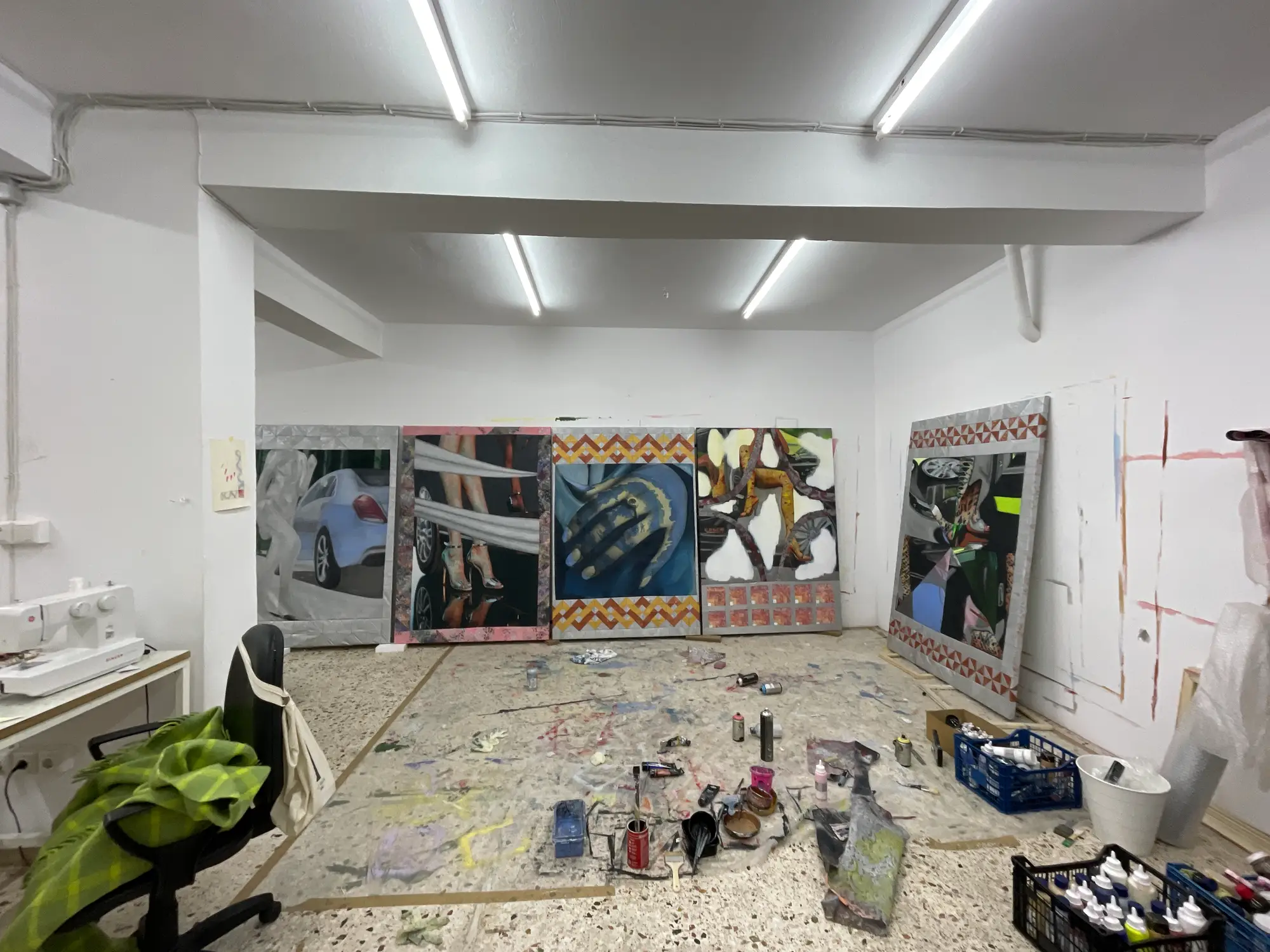Wide-angle view of Bety Krňanská’s Athens studio, showing multiple large paintings featuring cars and feminine motifs, with art supplies and sewing equipment scattered across the floor