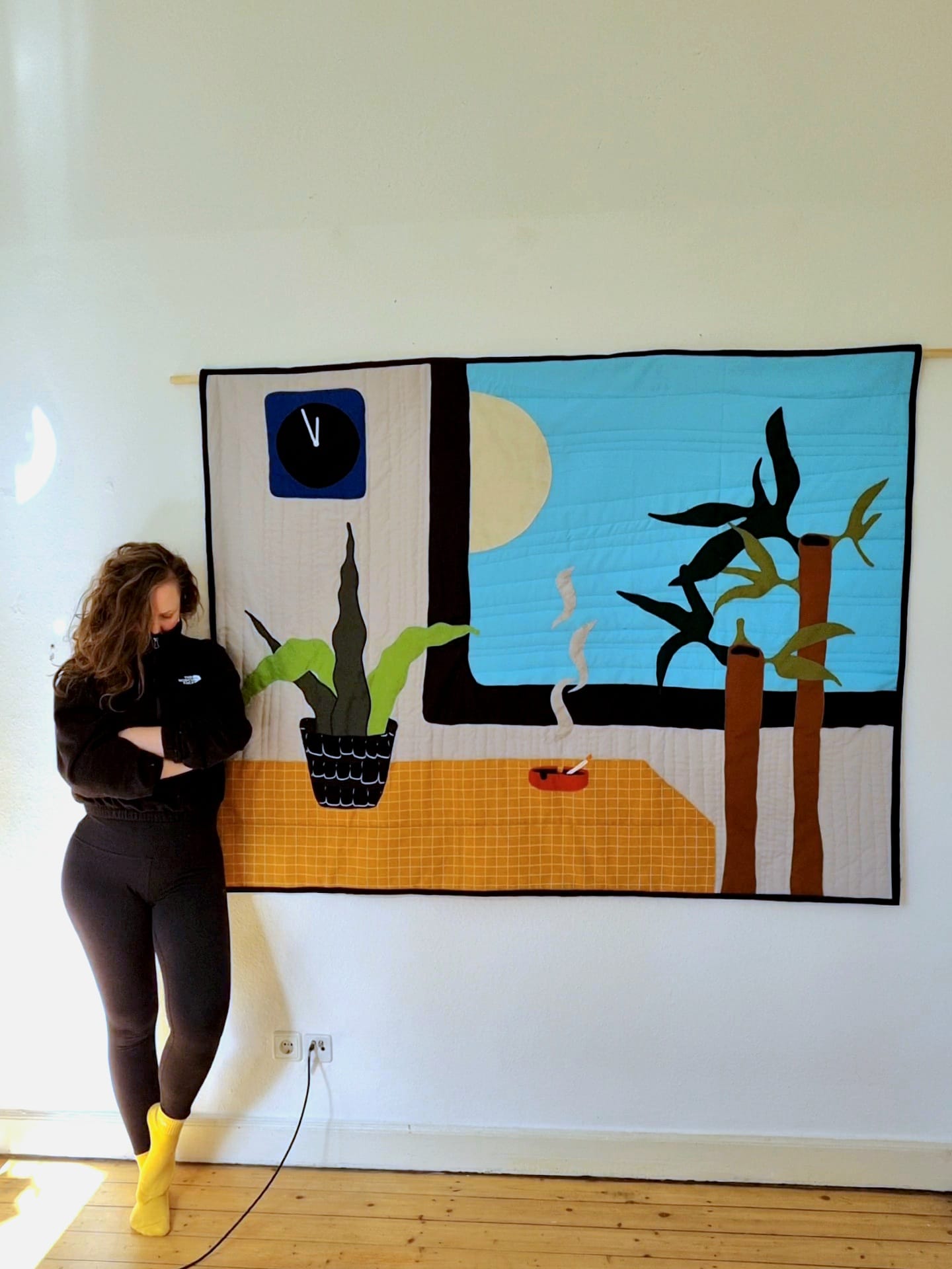 Artist Rosina Rosinski standing in front of her textile artwork Gutbürgerlich (2025), made from cotton and polyester, depicting a domestic interior scene with a window, plants, and a cigarette burning in an ashtray.