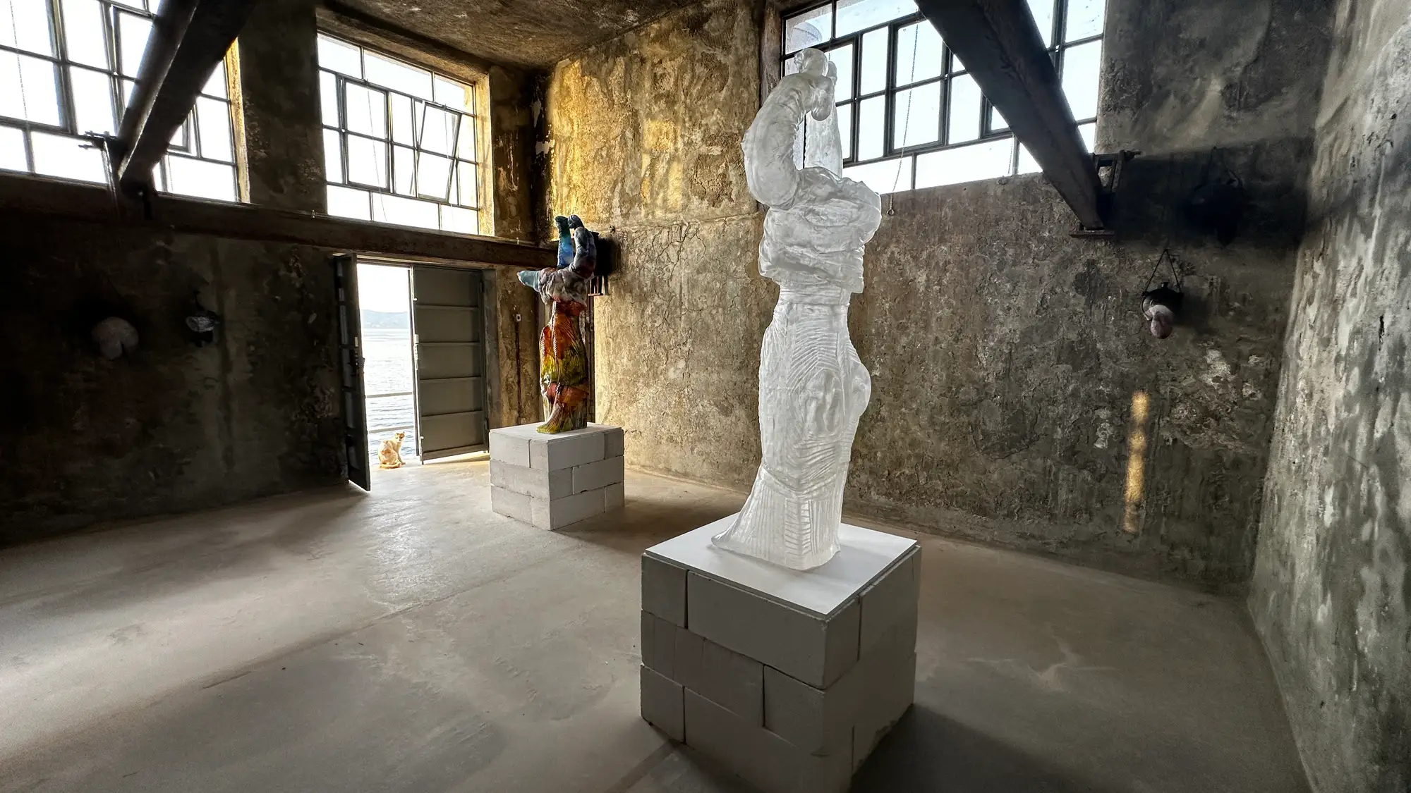 Andra Ursuța, “Apocalypse Now and Then,” installation view at DESTE Foundation Project Space (Slaughterhouse), Hydra, Greece, 2025—translucent cast-glass and colorful mixed-media sculptures on concrete-block plinths, sea visible through open doors
