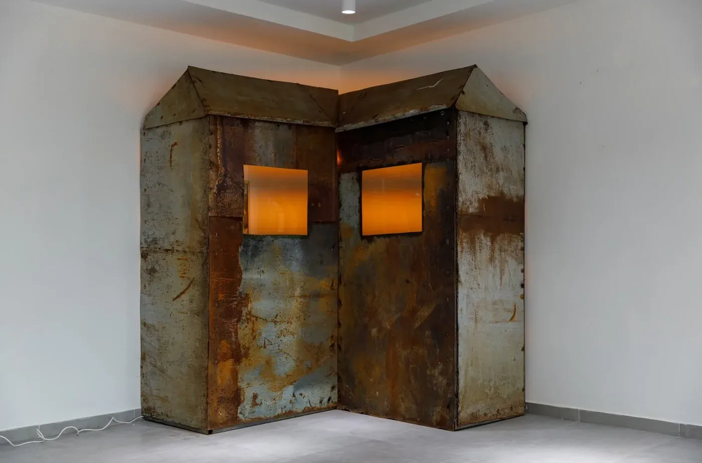 Mariam Akubardia, “River of Rust-Coloured Tears.” Rusted twin hut installation with amber-lit windows.
