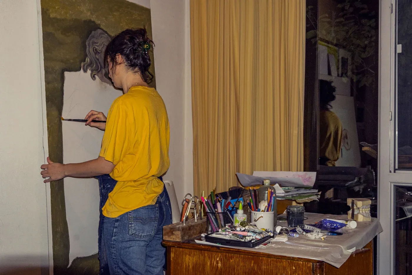 Mariam Akubardia working on a painting at an easel, table with brushes and tools nearby.