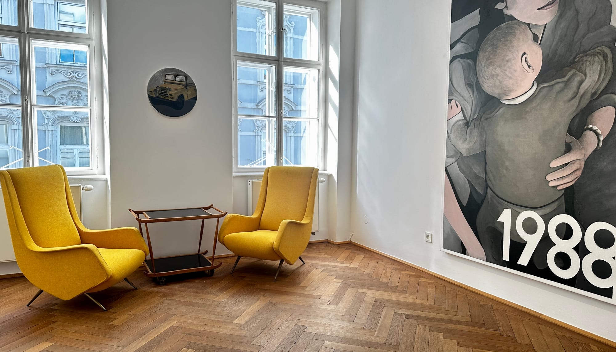 Alt text: Seating area at Klubhaus with a round Jeep painting on the wall and the edge of the mother-and-child painting from “1996.”