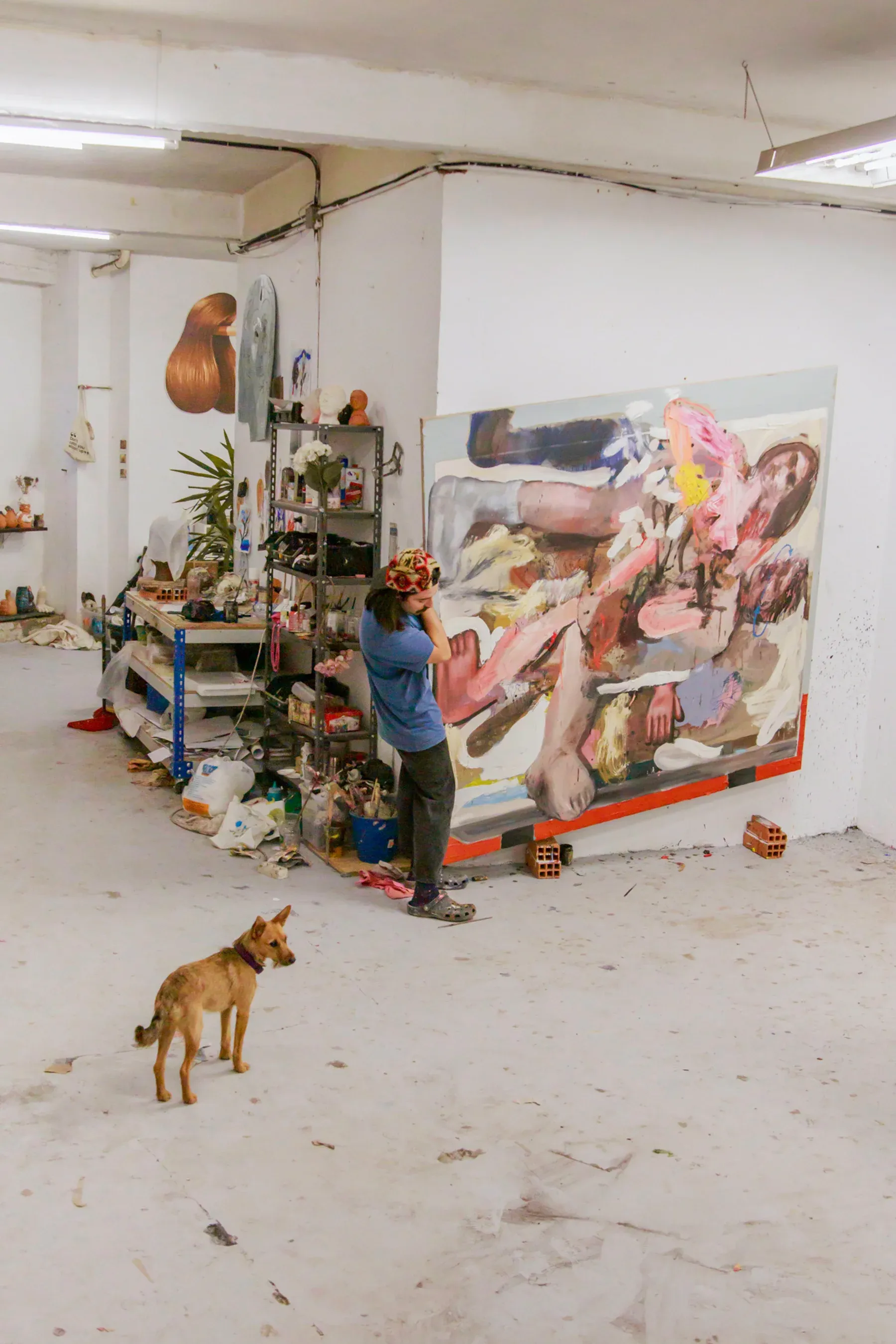 Manuel García Fernández in his studio with a dog standing nearby, surrounded by paint materials and a large figurative painting. Photo by Juan García-Mendoza.
