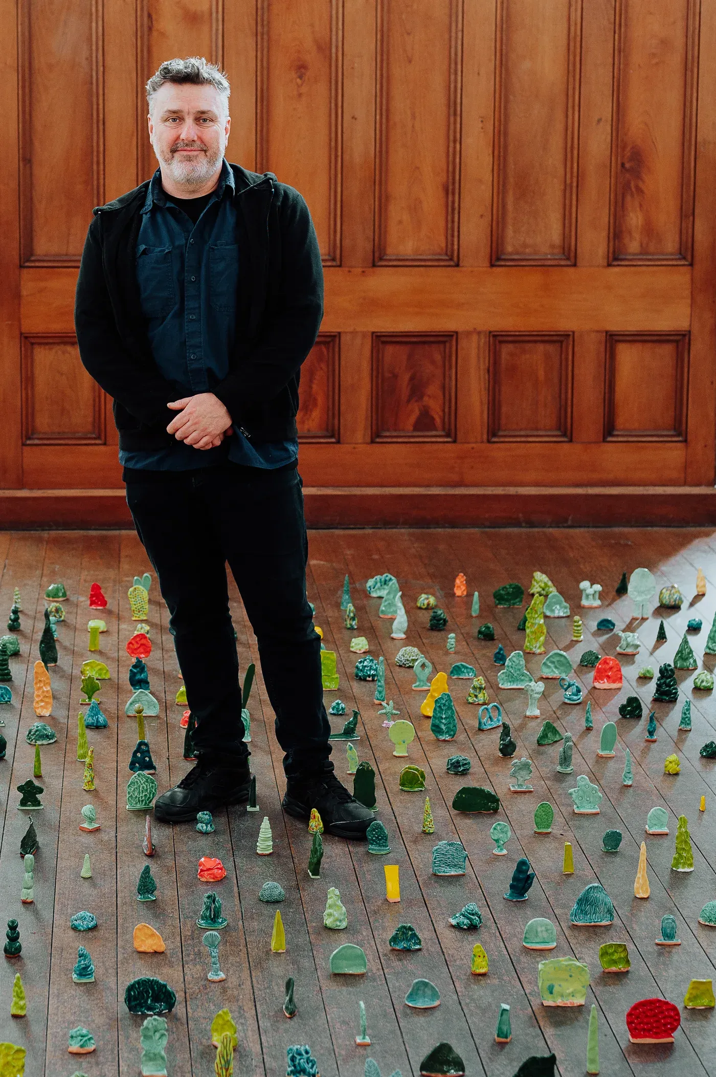 Glenn Barkley standing in his studio, surrounded by a garden full of small colorful ceramic works arranged on a wooden floor, photographed by Berry NSW.