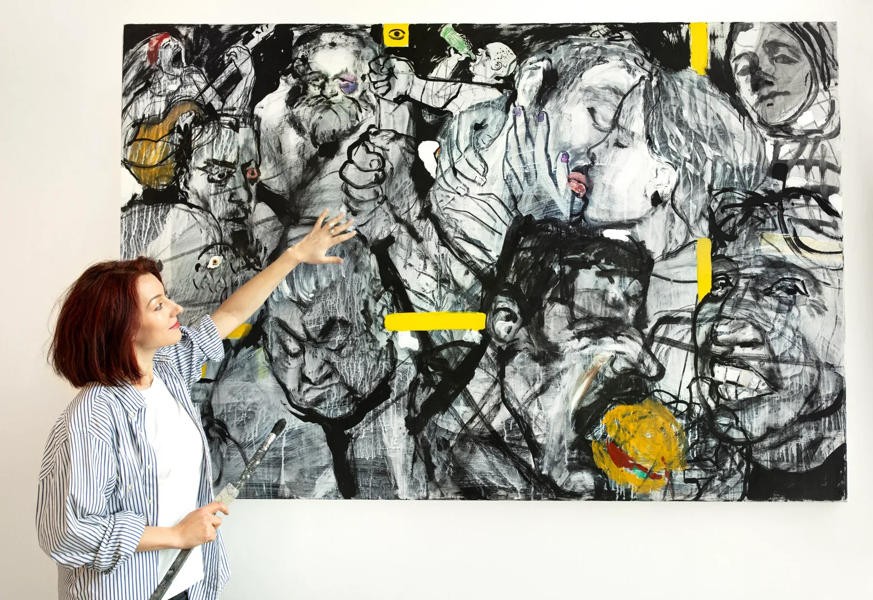 Artist Maria Naidyonova standing beside her large mixed-media painting Metro, a dense composition of expressive overlapping faces and figures in black, white, and yellow accents.