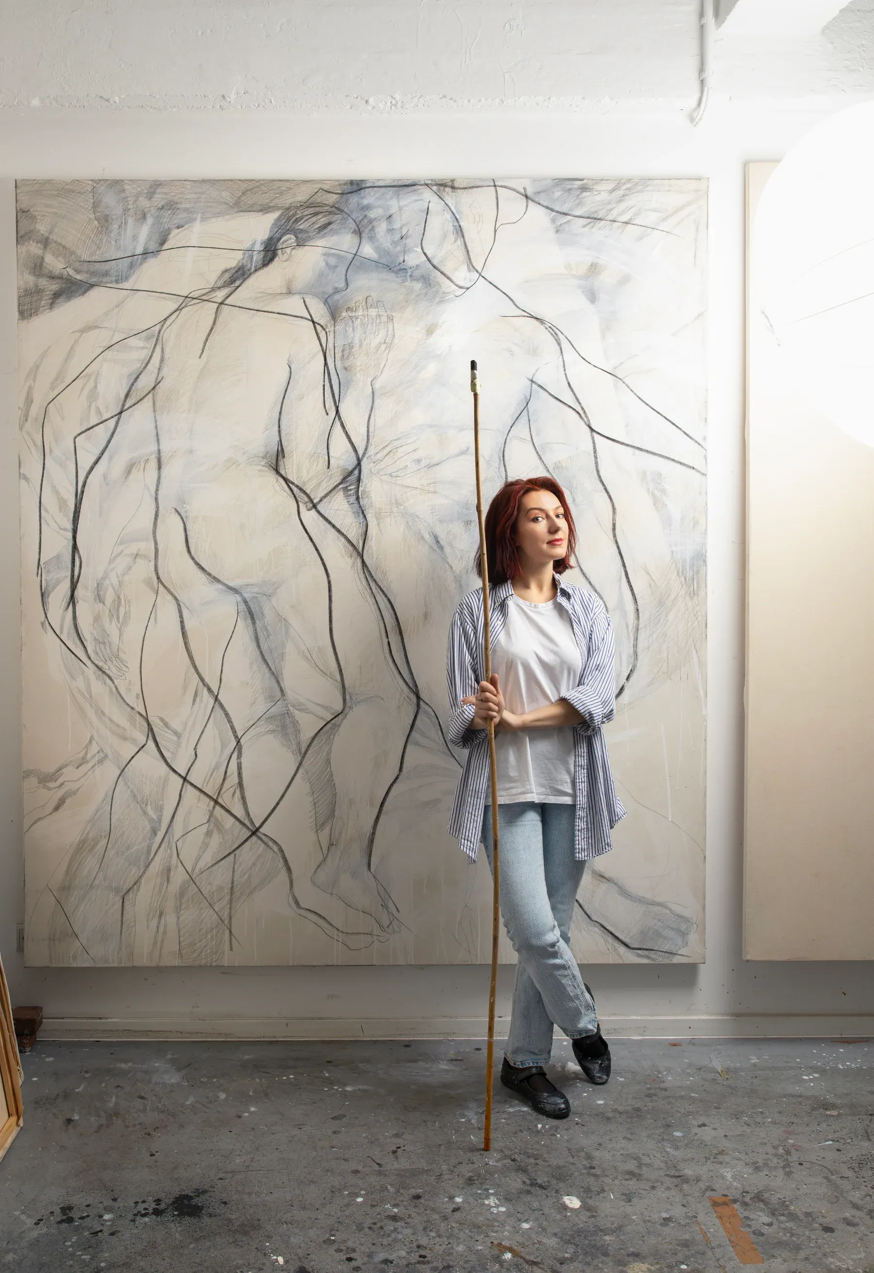 Maria Naidyonova standing in her studio holding a long stick, positioned in front of a large gestural figure drawing on canvas.