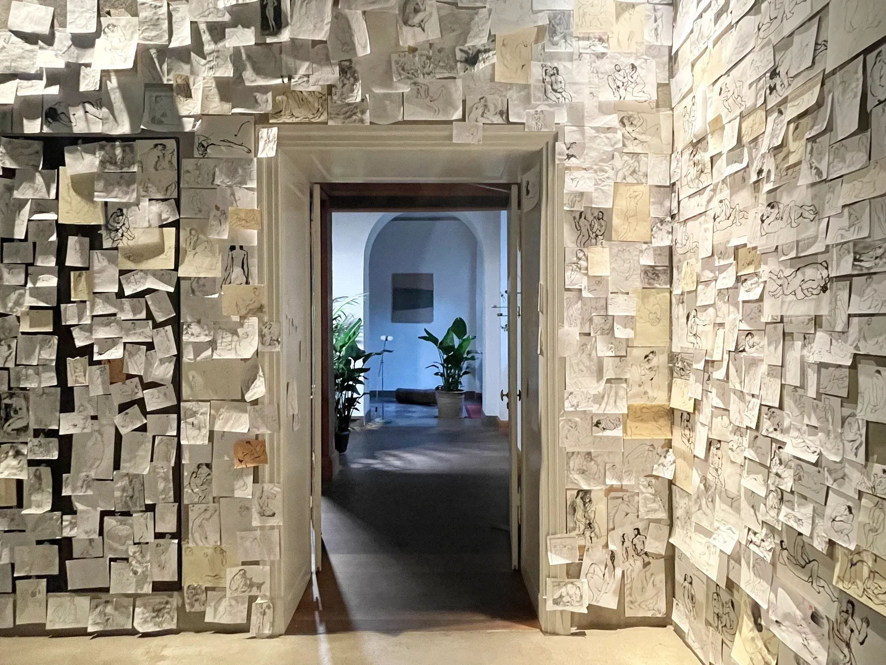 A gallery hallway covered floor-to-ceiling with small figurative drawings by Maria Naidyonova, creating a dense collage-like installation around a central doorway.