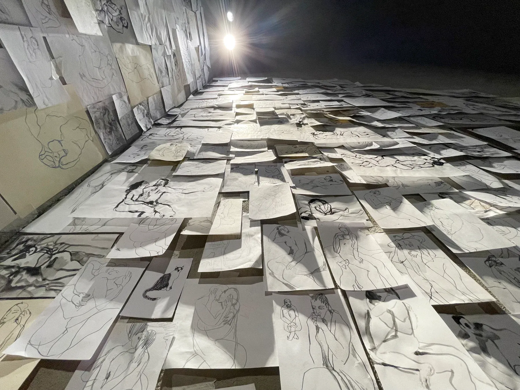 An immersive installation by Maria Naidyonova showing hundreds of drawings mounted on a tall gallery wall, viewed from below toward a ceiling light.