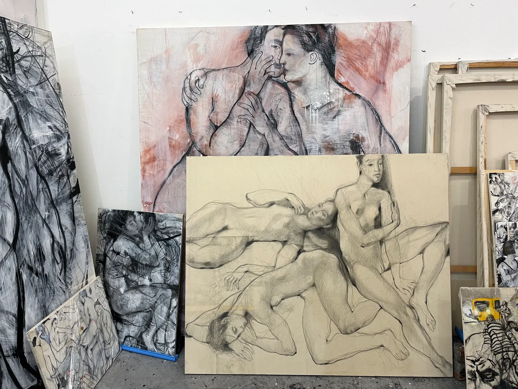 A studio corner with multiple large figurative paintings by Maria Naidyonova leaning against the wall, showing intertwined bodies drawn in charcoal, graphite, and paint.