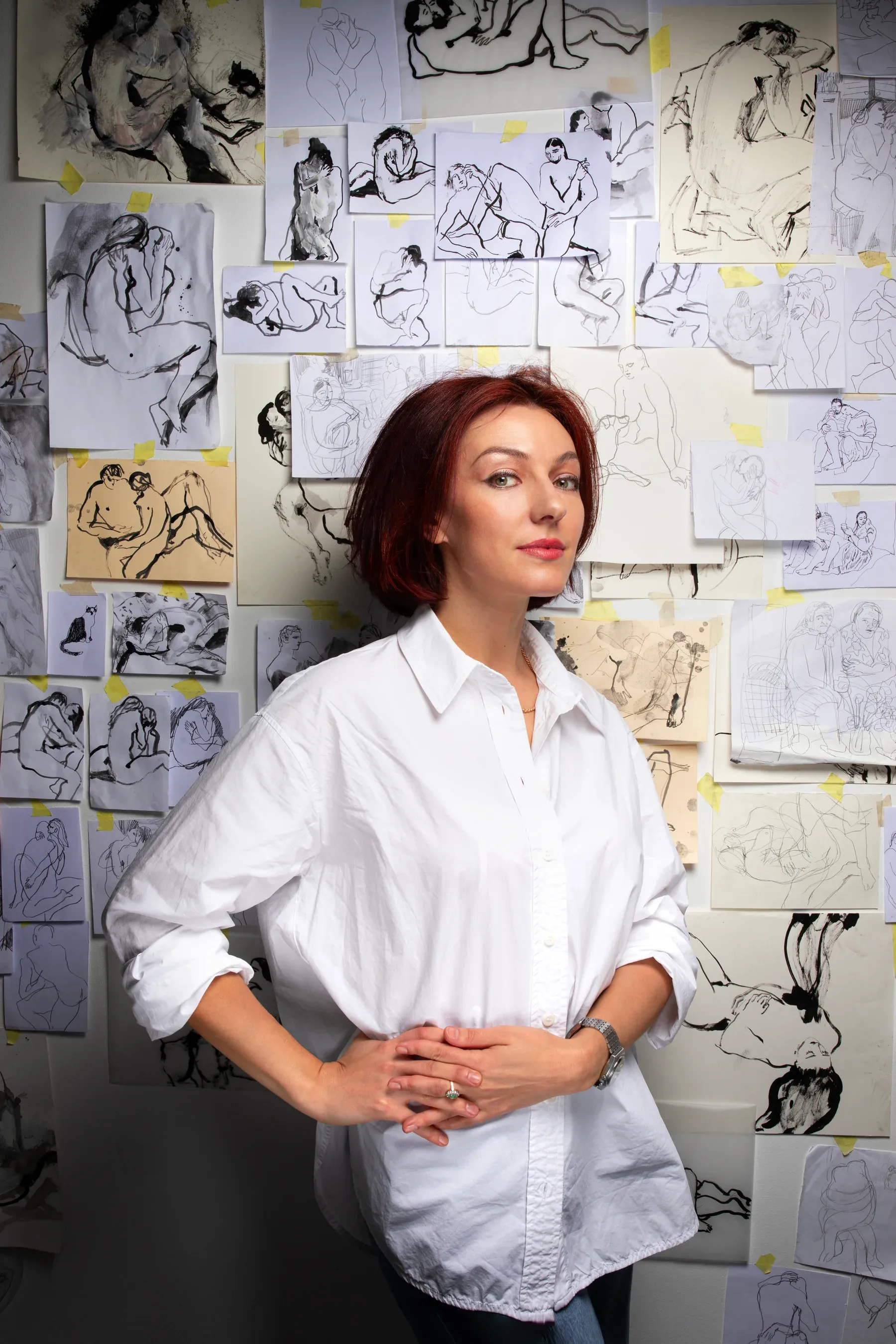 Artist Maria Naidyonova standing in front of a studio wall filled with taped drawings of nude figures, wearing a white shirt and looking directly toward the camera.