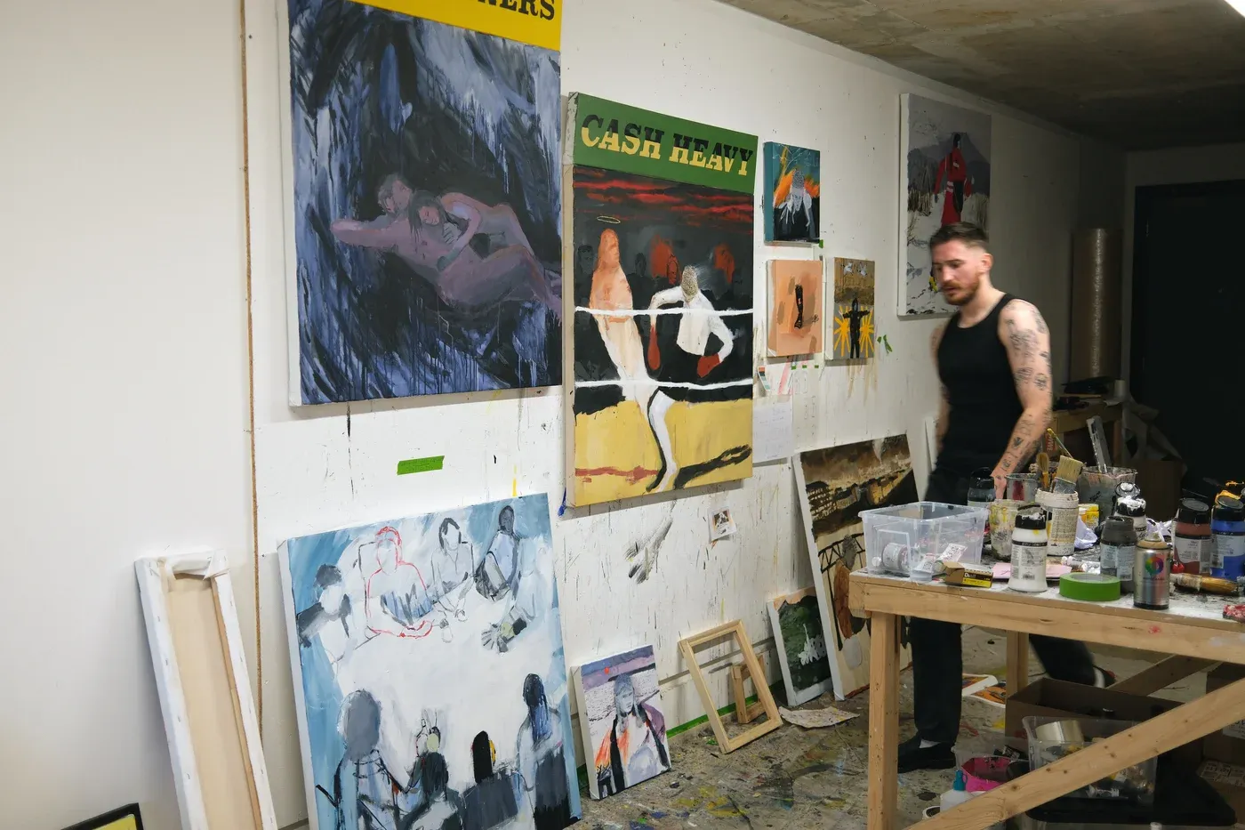 Studio view showing artist Billy Bagilhole standing beside several paintings on the wall, including Cash Heavy, surrounded by paint jars, brushes, and unfinished canvases.
