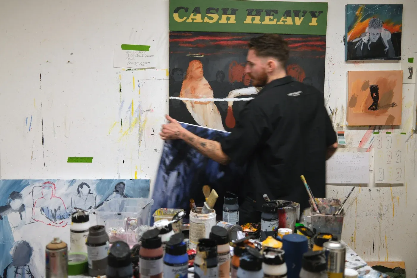 Artist Billy Bagilhole in his studio, holding a canvas among paint jars and brushes, with several of his works, including Cash Heavy, visible on the wall.