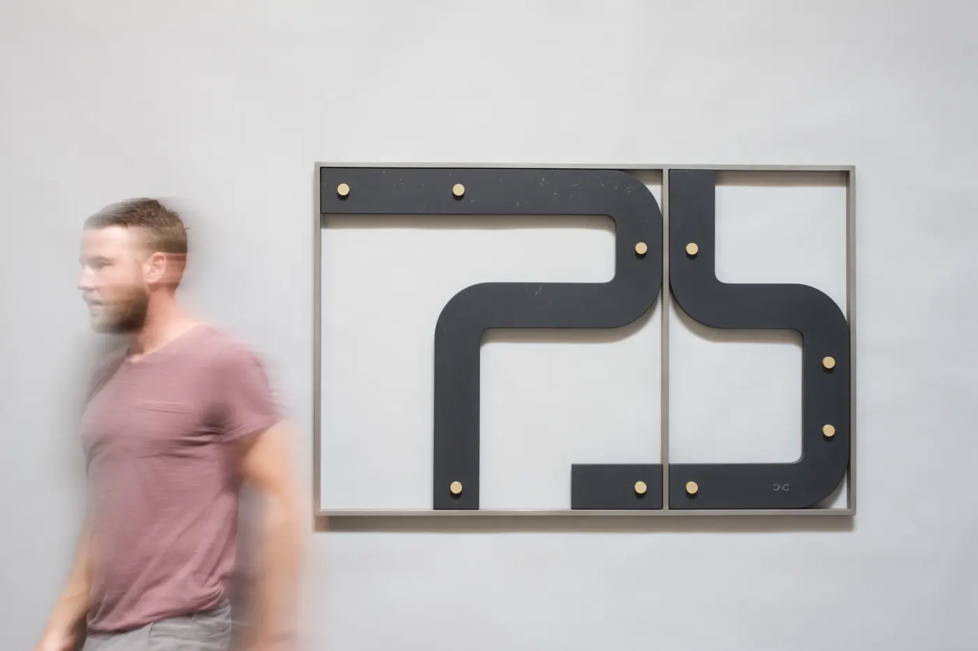 Artist Chris Cassimatis walking past his wall sculpture Construct No. 7, made of slate, stainless steel and brass, with curved dark elements mounted in a metal frame. Image Courtesy of the Artist