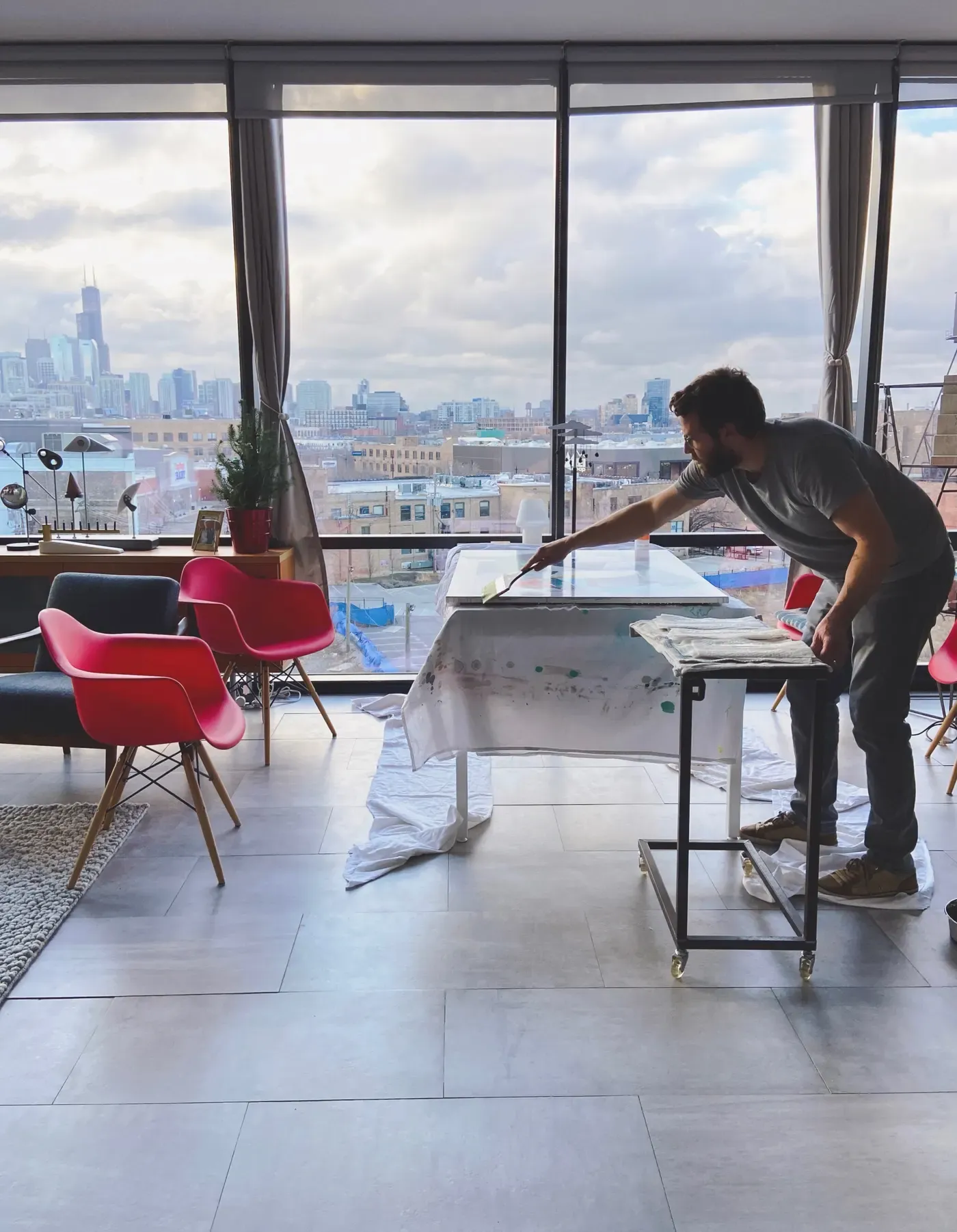 Chris Cassimatis working on a painting in his Chicago kitchen table studio with city skyline in the background, showing his early workspace setup from 2009 to 2022.