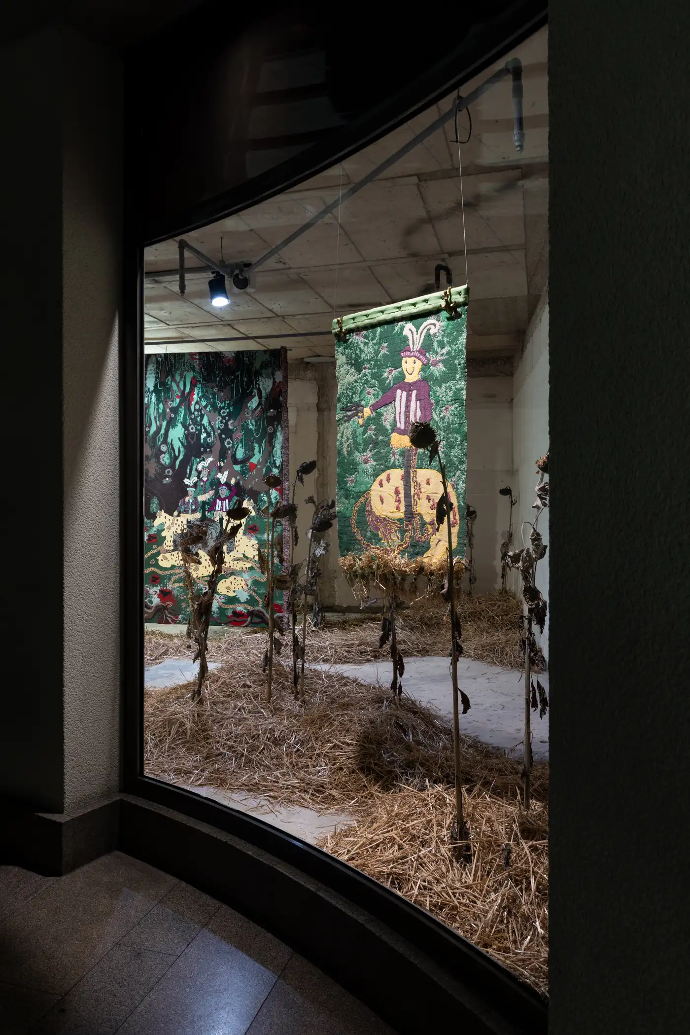 Nighttime view of Olivia Rode Hvass’s installation HUNT(ED), seen through illuminated windows with woven tapestries and dried sunflowers. Photographed by Patty Neu