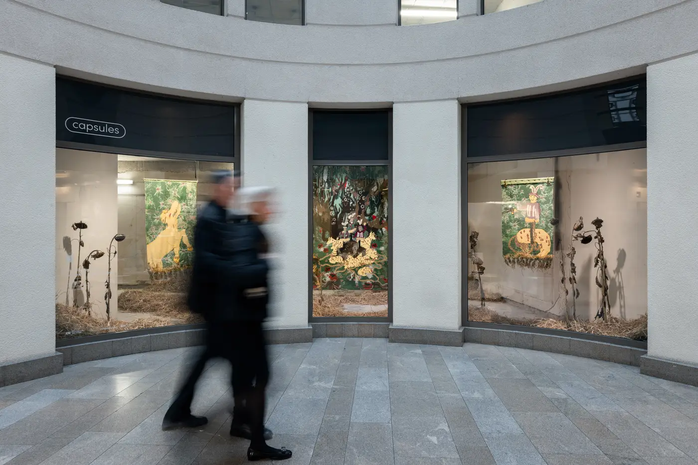 Street view of Capsules at Luxembourg Art Week showing Olivia Rode Hvass’s installation HUNT(ED) through three illuminated display windows with woven tapestries and dried sunflowers. Two blurred pedestrians walk past. Photographed by Patty Neu.