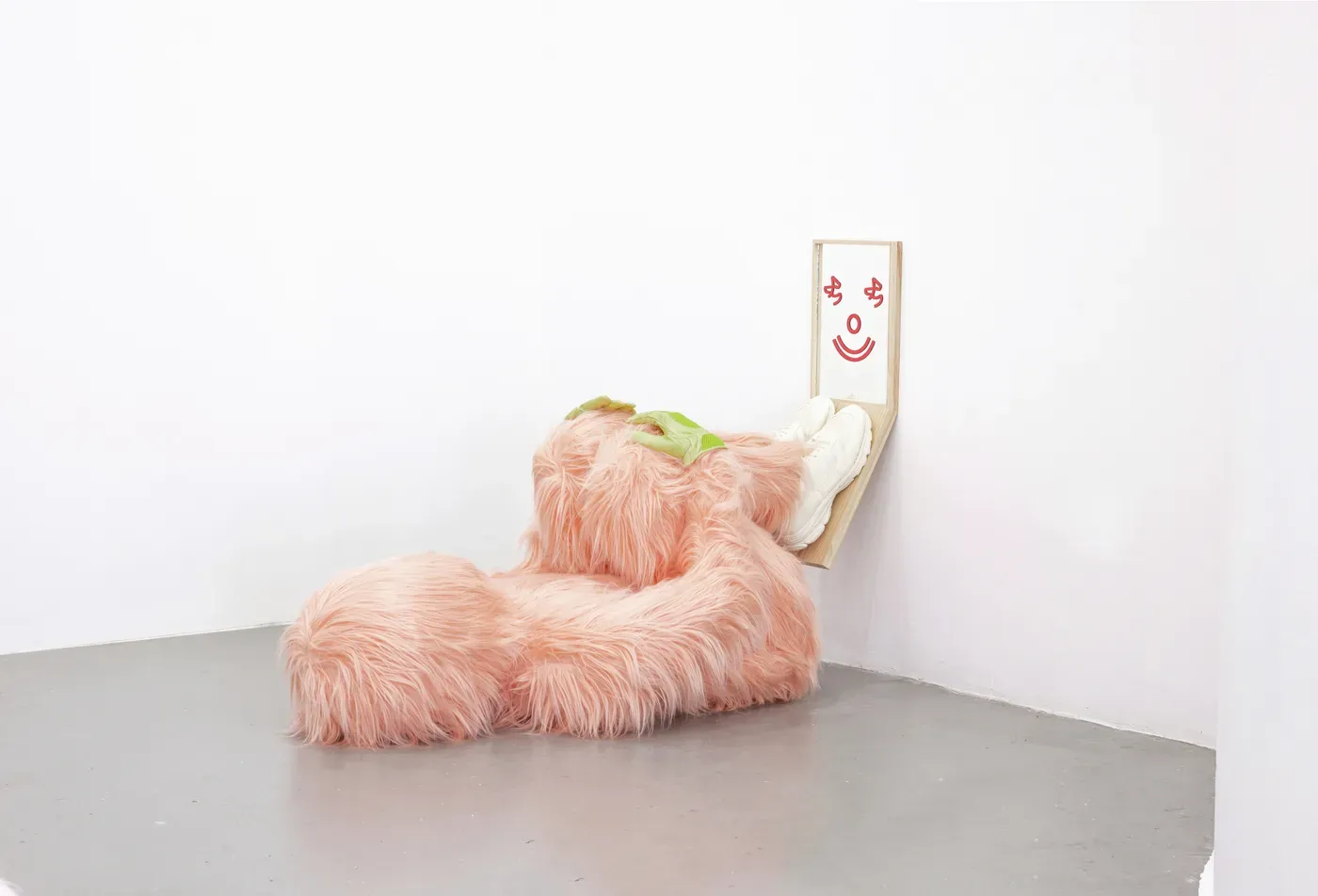 Sculpture Health Coach by Grip Face. A pink furry humanoid form with white sneakers leans against a wall featuring a smiling red face painted on wood. Made from cloth, resin, fibreglass, and synthetic leather. Exhibited at @scgallery_bilbao