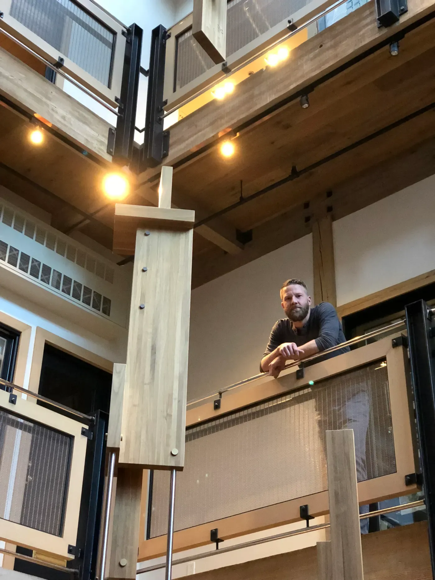 Artist Chris Cassimatis standing on an upper level next to a tall wooden sculptural installation in Aspen, shown in an architectural interior with warm lighting. Image Courtesy of the Artist.