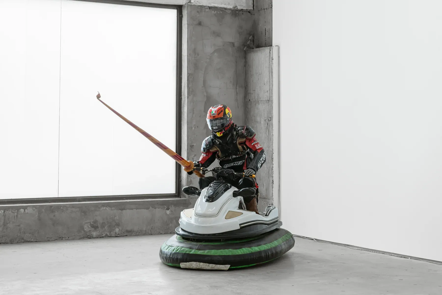 Armored figure riding a modified bumper car while holding a long lance, positioned in a corner of a bright concrete gallery.