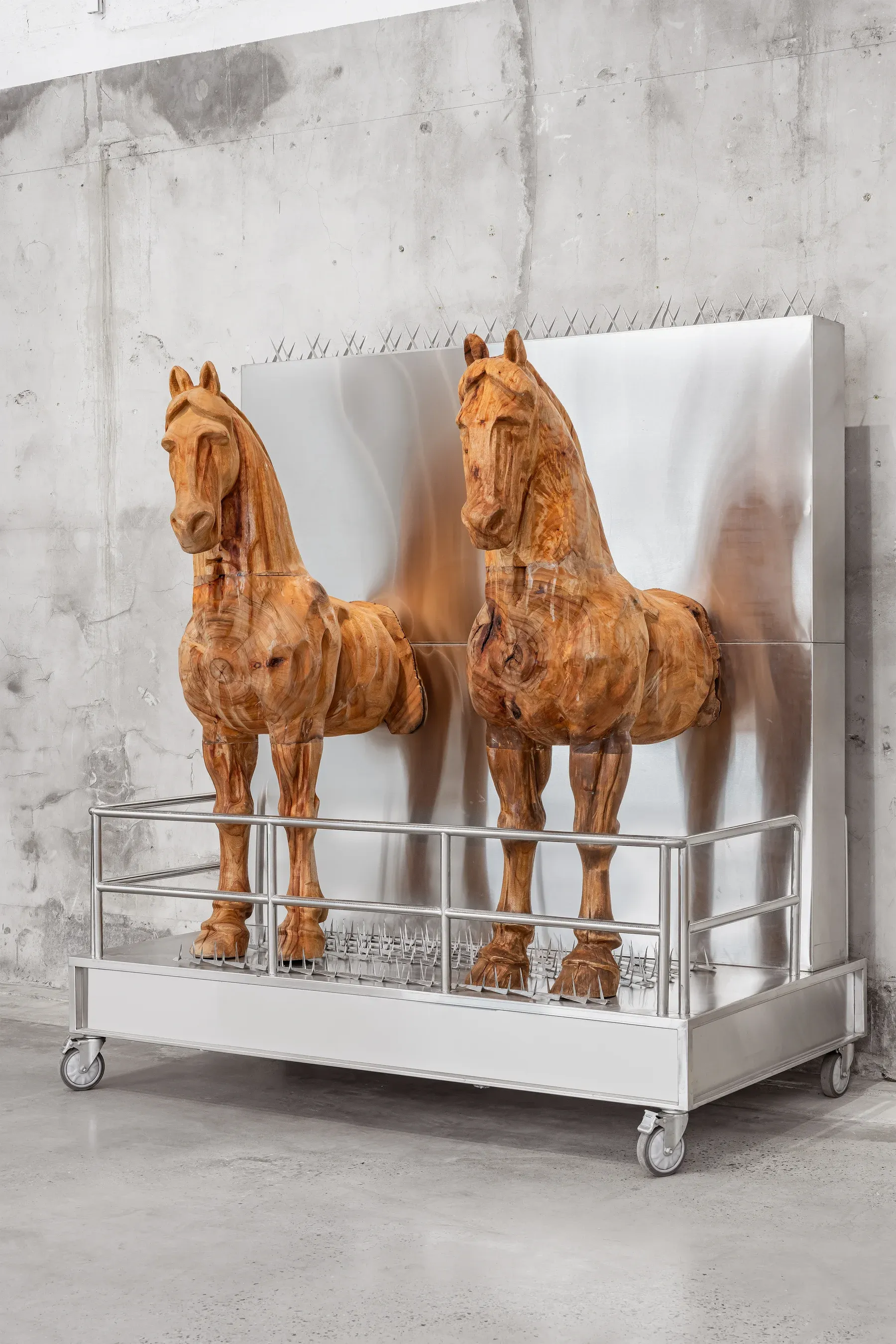 Two carved wooden horse sculptures presented on a wheeled metal platform with a reflective steel backdrop.