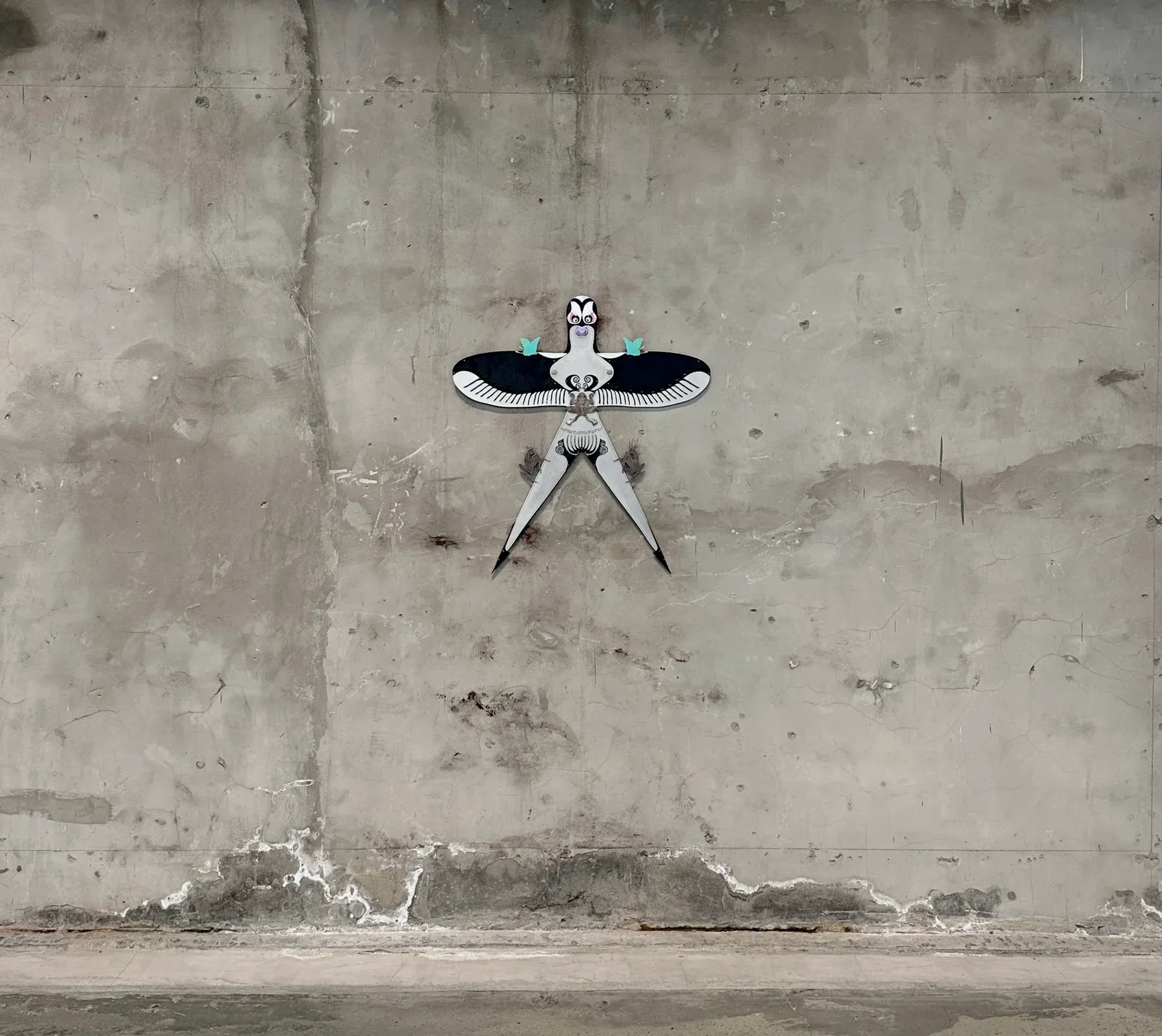 Wall-mounted sculpture resembling a stylized bird figure with extended wings and long legs installed on a weathered concrete wall.