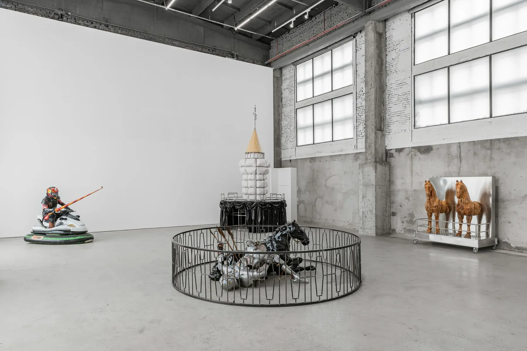 Gallery installation featuring a padded tower, a fallen metallic horse sculpture inside a circular barrier, and wooden horses against a reflective panel.