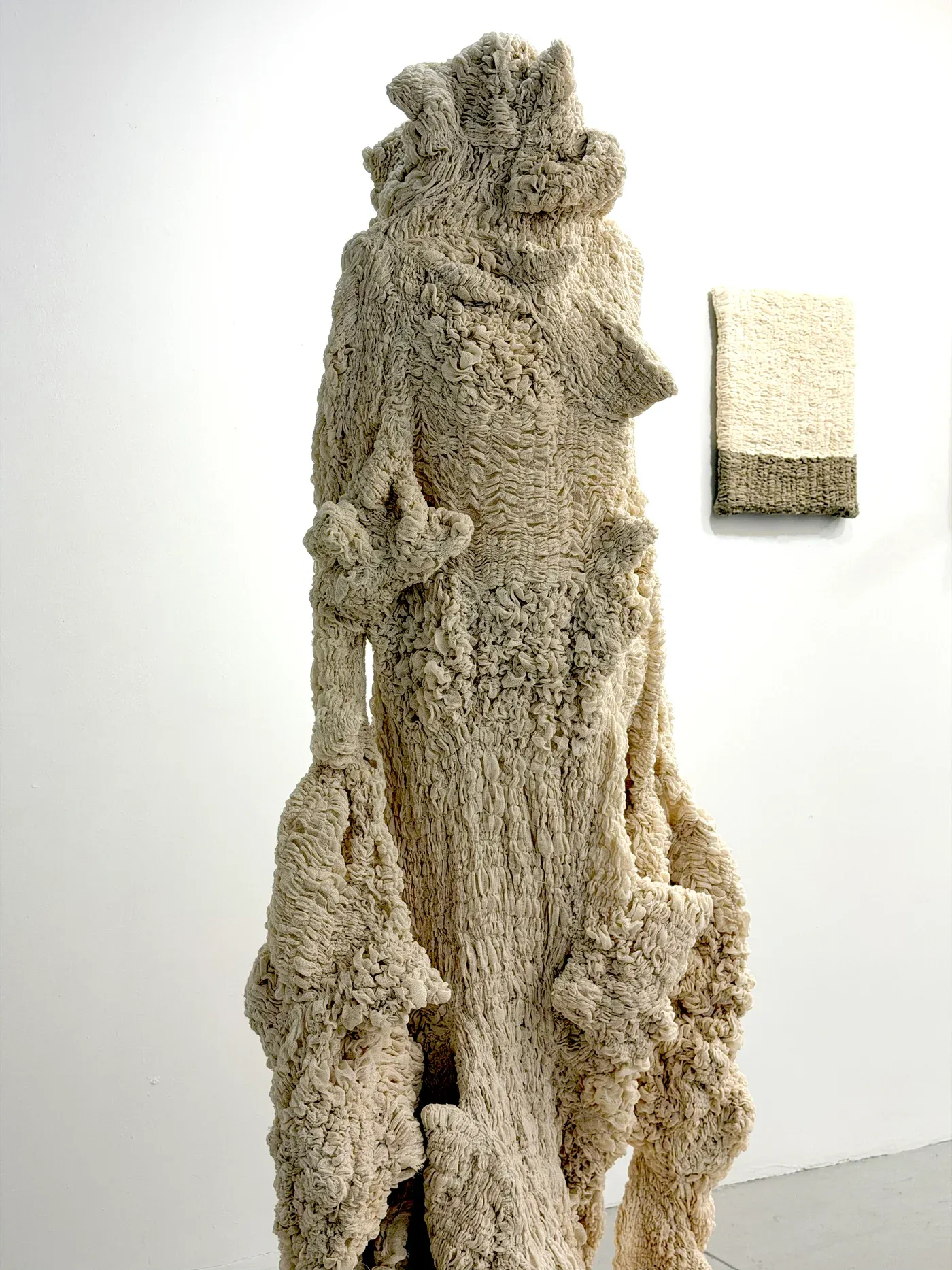 Yali Romagoza, installation view at The Elizabeth Foundation for the Arts, NY, showing a tall linen and thread sculpture with intricate folds and textures beside a smaller wall-mounted fiber panel.