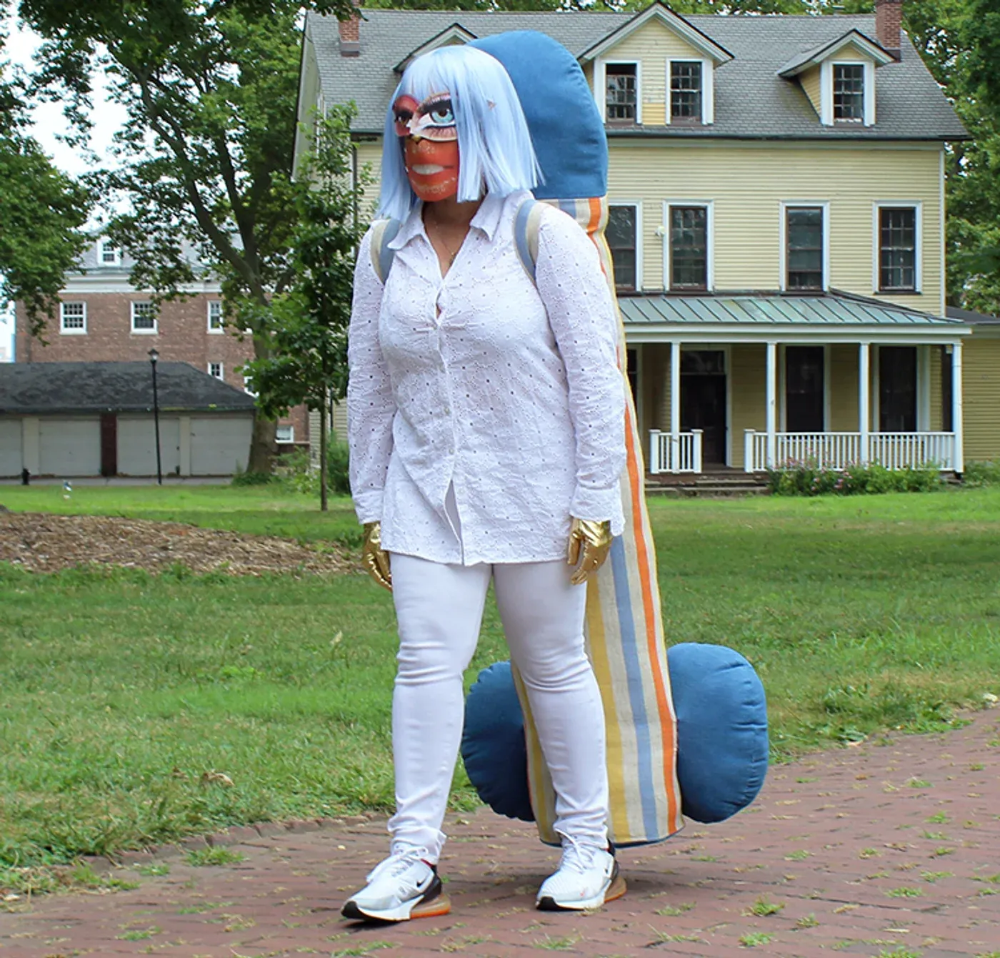 Yali Romagoza performing A Cross to Bear (2024), wearing a blue wig, mask, white clothing, and gold gloves while carrying a large striped soft sculpture outdoors during Rebirth of a Body in New York.