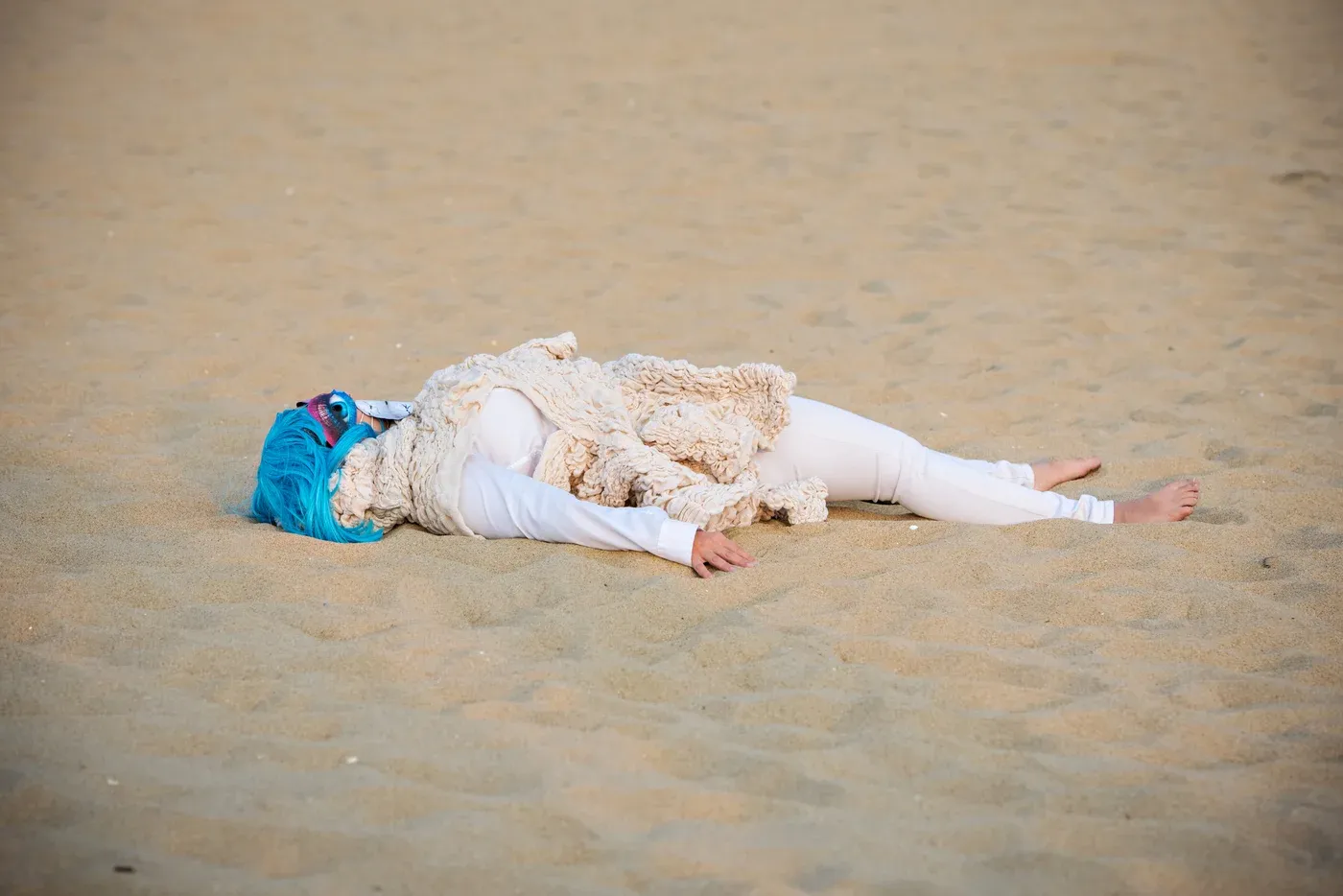 Yali Romagoza lying on sand during her performance Meditating Ways to Escape Body Trauma (The Healing Dance) (2023), wearing a linen textile sculpture, white clothing, and a blue wig at Asbury Park Beach, NJ.