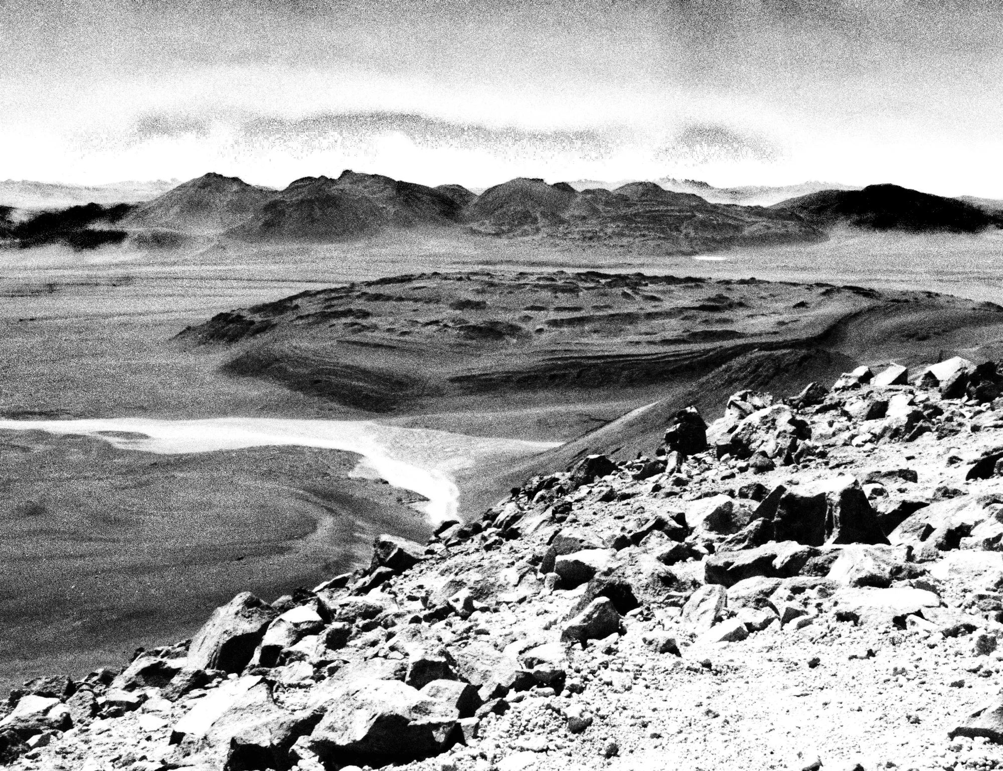 Black-and-white desert landscape photograph from Emergent Phenomenon by Nadine Karl, depicting layered terrain and distant mountain ranges.