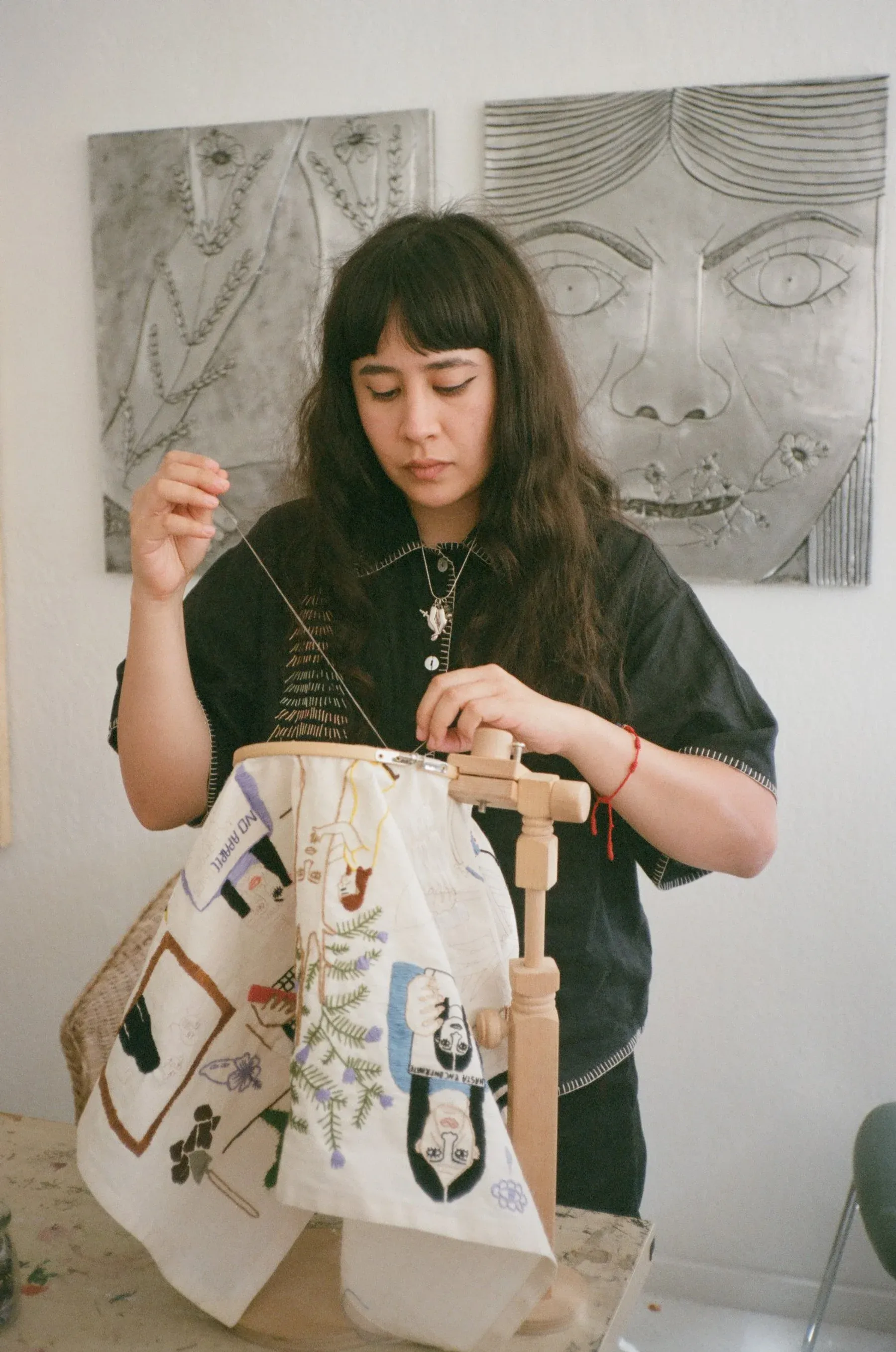 Artist sitting in her studio stitching embroidered imagery onto fabric, with metal relief portraits hanging behind her.