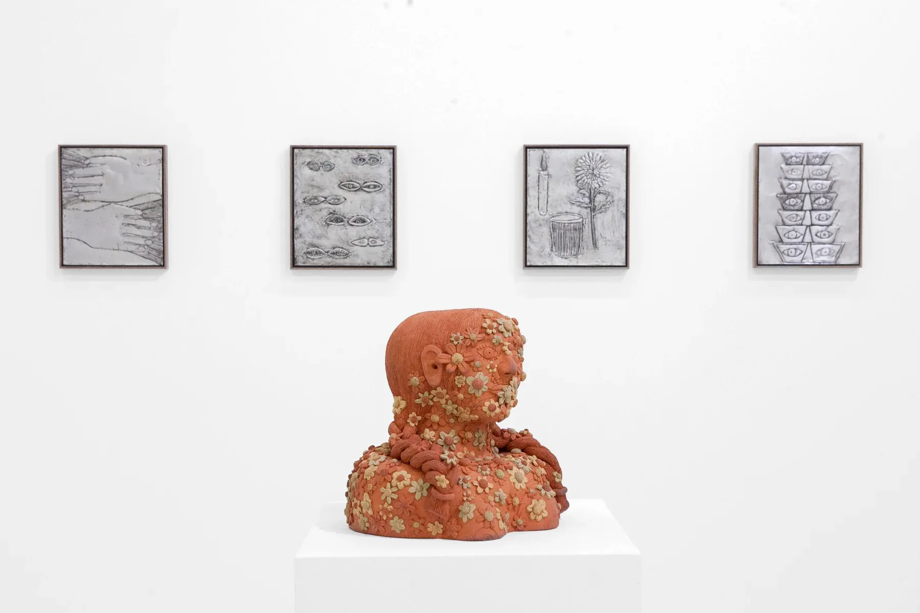Terracotta sculpture covered in textured floral elements placed on a pedestal, with framed drawings on the white gallery wall behind.