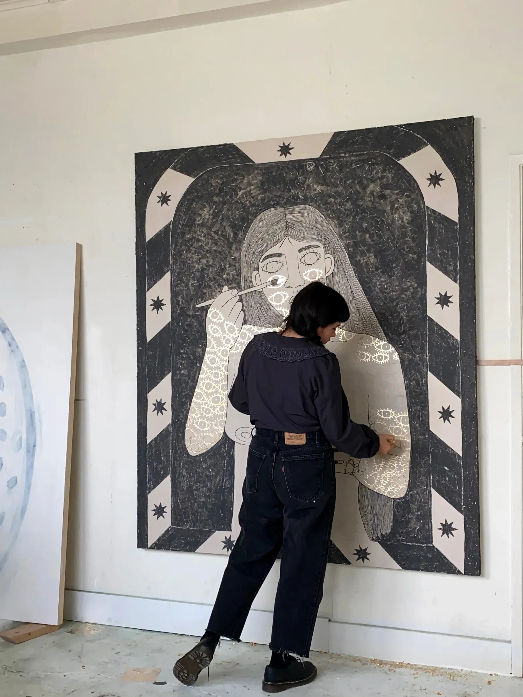 Artist standing in front of a large black-and-white canvas, painting floral patterns onto a figure’s dress.