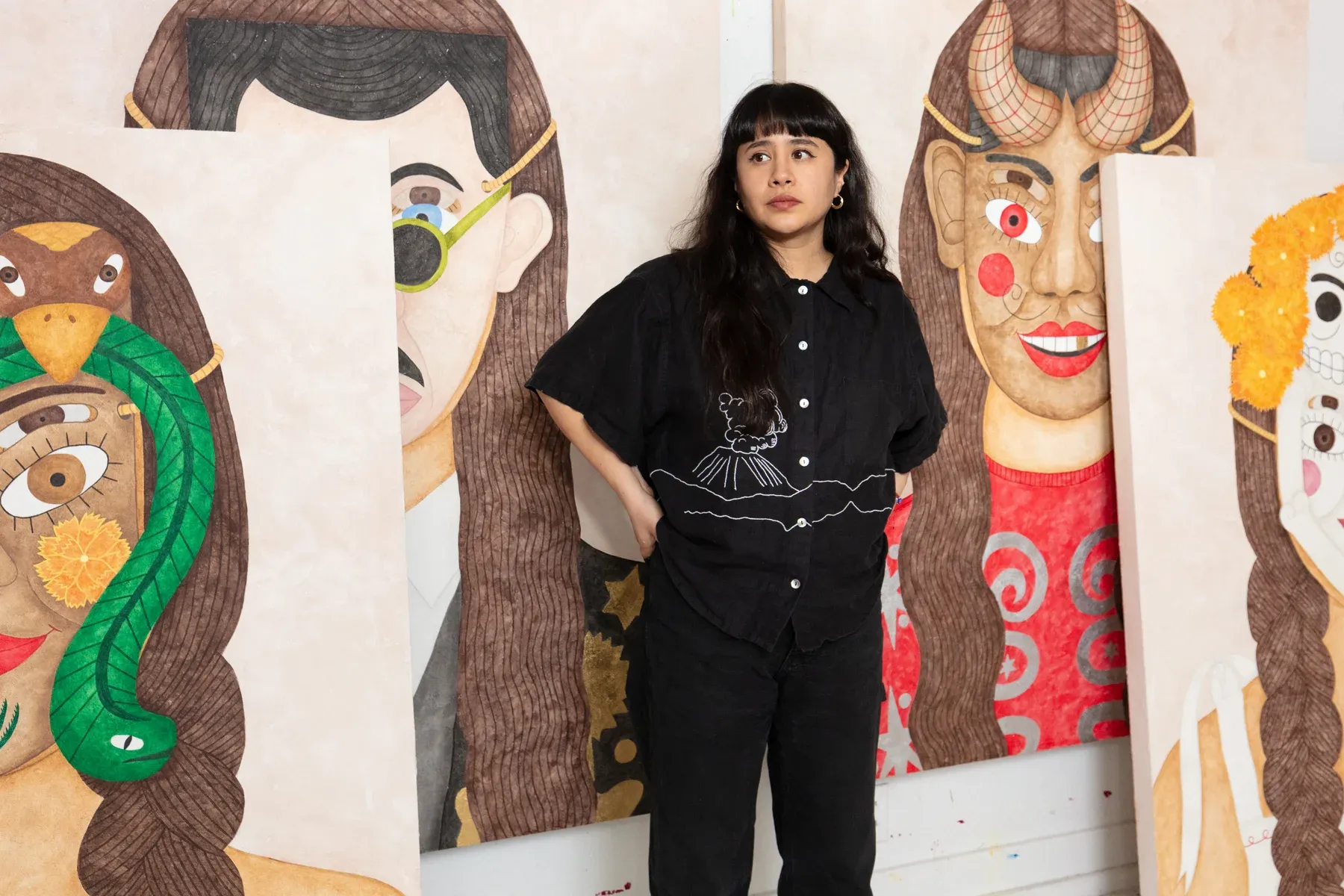 Liz Hernandez standing in a studio surrounded by large painted portraits featuring mask-like composite faces.