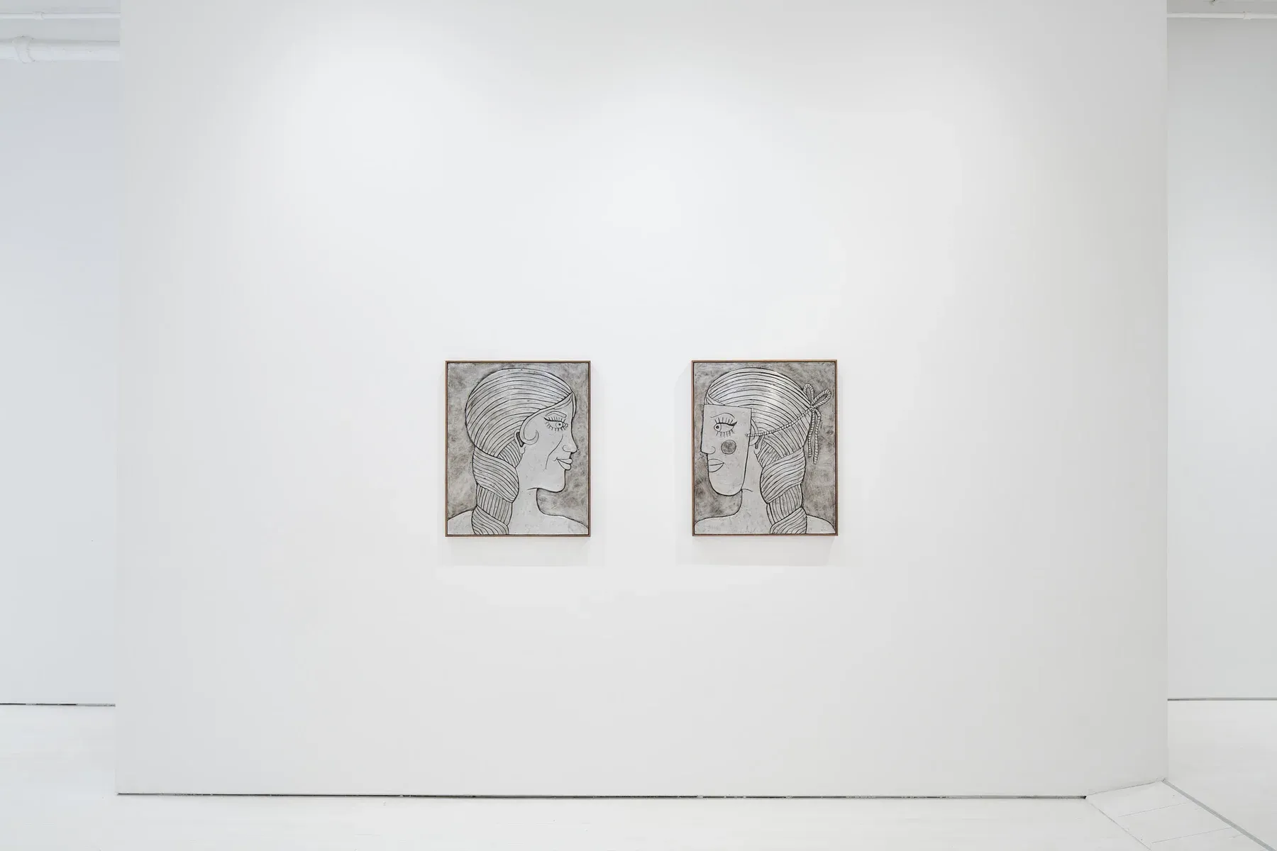 Two framed black-and-white portraits with mask-like profiles hanging side by side on a white gallery wall.