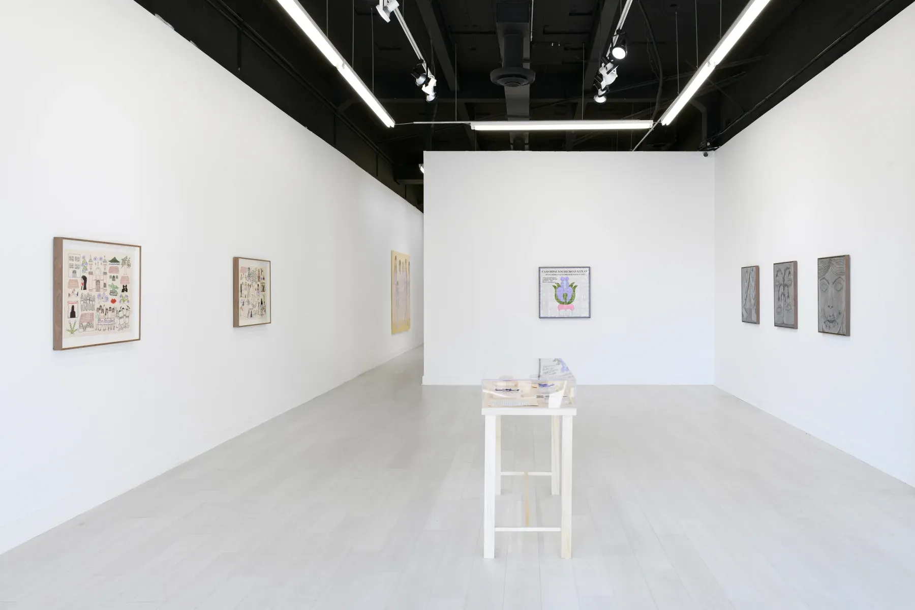 Minimal white-walled gallery with framed works hung along both sides and a small table in the center displaying drawings and objects.