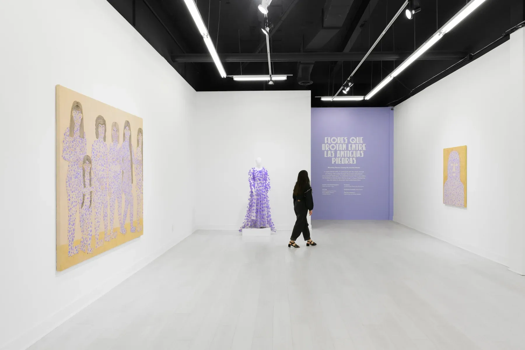 Gallery view showing lavender-toned paintings, a dress sculpture, and a visitor walking toward a purple wall text.