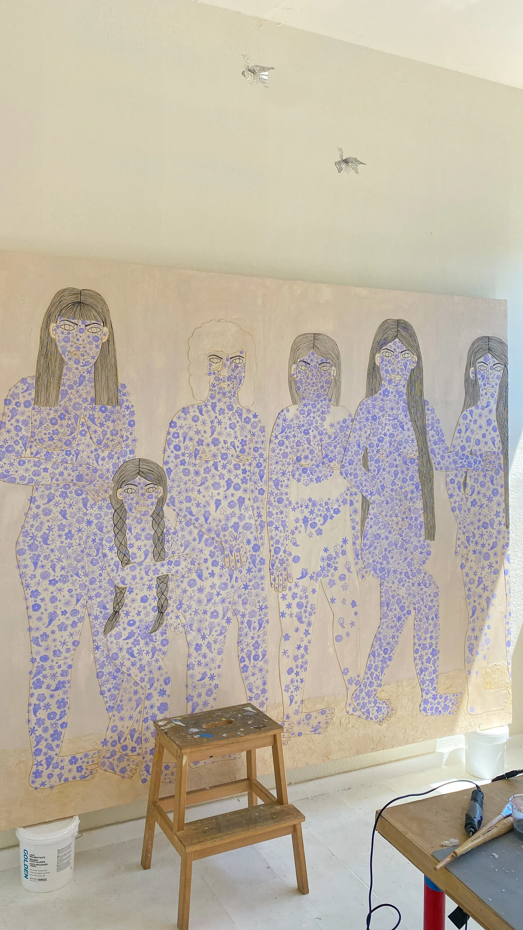 Large canvas showing seven figures outlined and filled with small purple flowers, with a stool and painting tools in front.