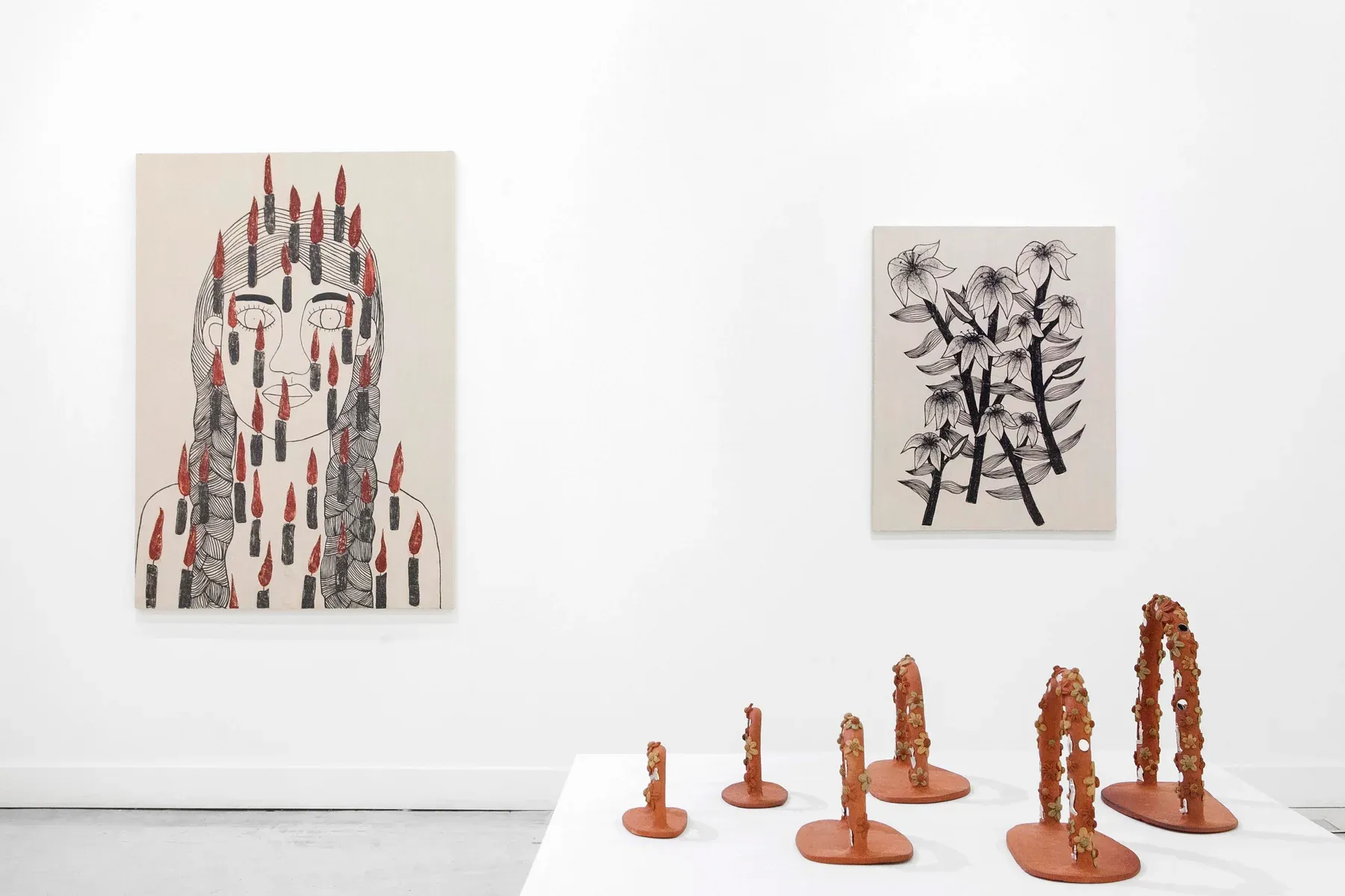 Sculptures shaped like vertical organic forms in terracotta placed in front of two paintings depicting braided figures and botanical drawings.