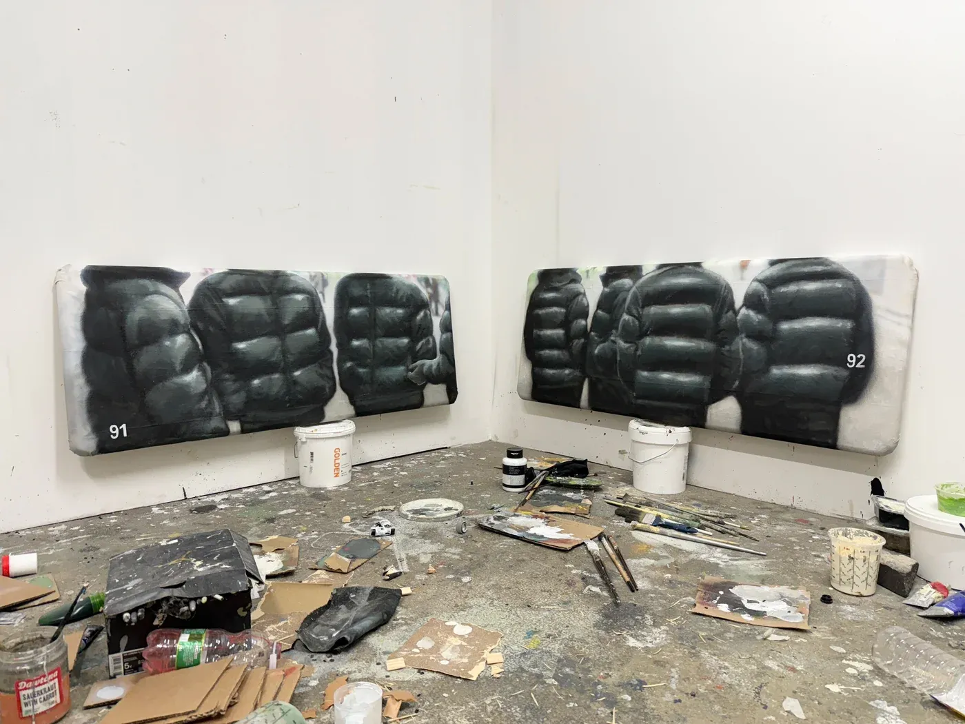 Yutaro Inagaki, Two long horizontal puffer jacket paintings, marked 91 and 92, resting on paint buckets in a cluttered studio corner.