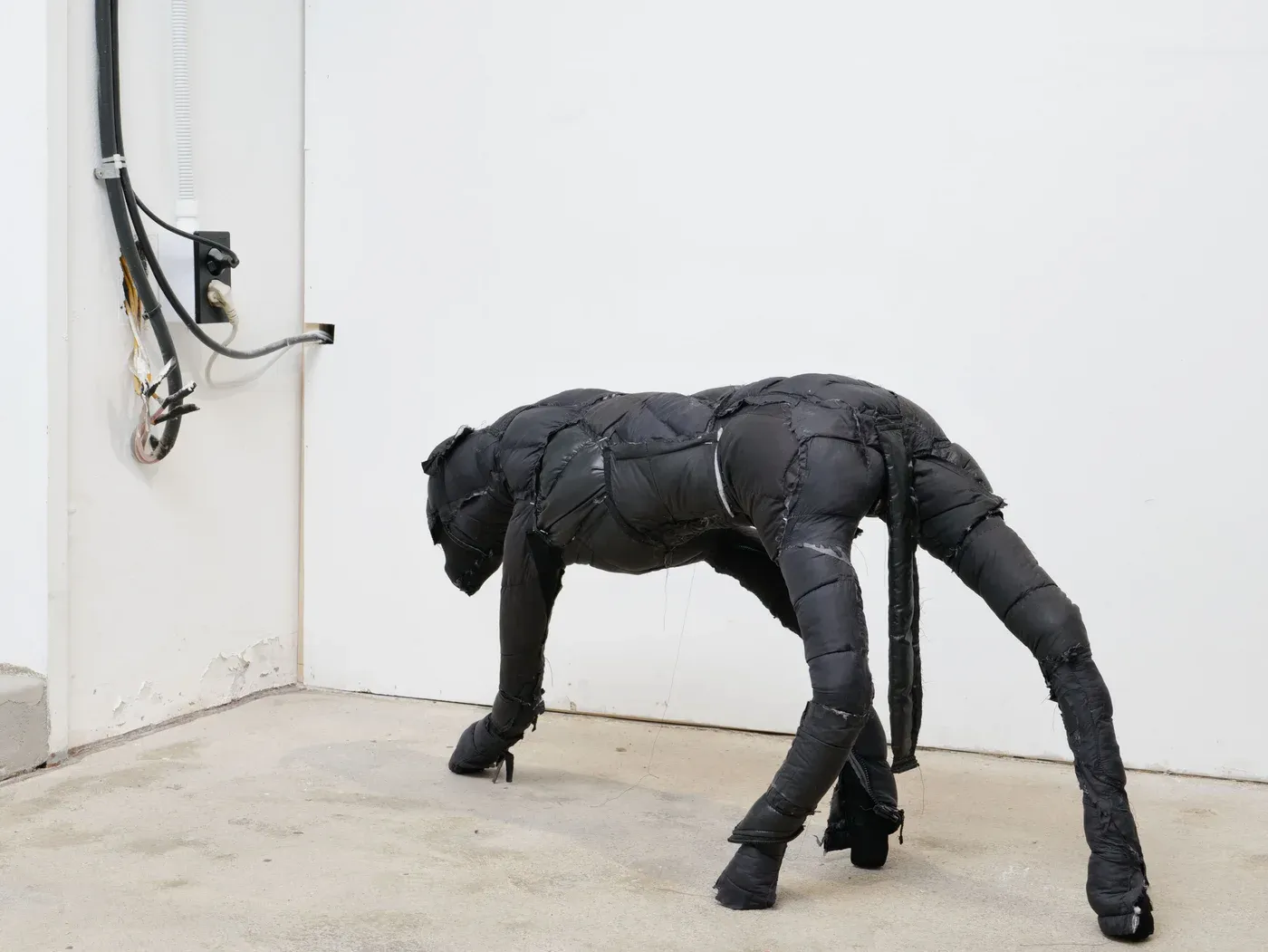 Yutaro Inagaki, Full view of the Dog B sculpture, a black puffer jacket covered dog form standing on four legs in a white walled space.
