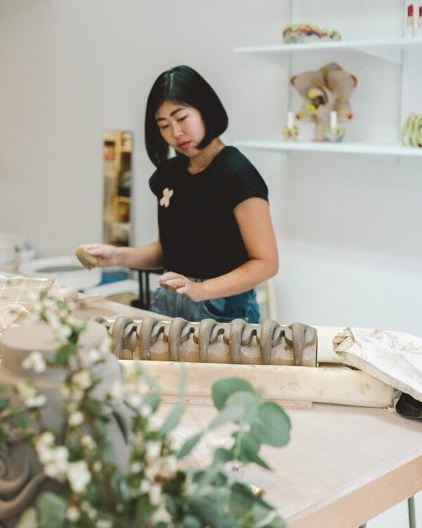 Artist Eri Maeda shaping a ceramic piece in her studio, wearing a black shirt and working at a long table with unfinished stoneware forms.