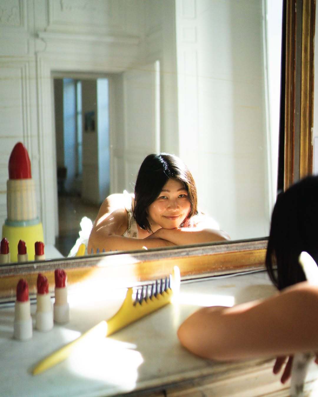 Eri Maeda leaning on a mirror ledge, reflected beside her ceramic beauty tool sculptures including Monster Comb and lipstick forms. Soft morning light, dress by Cecilie Bahnsen.