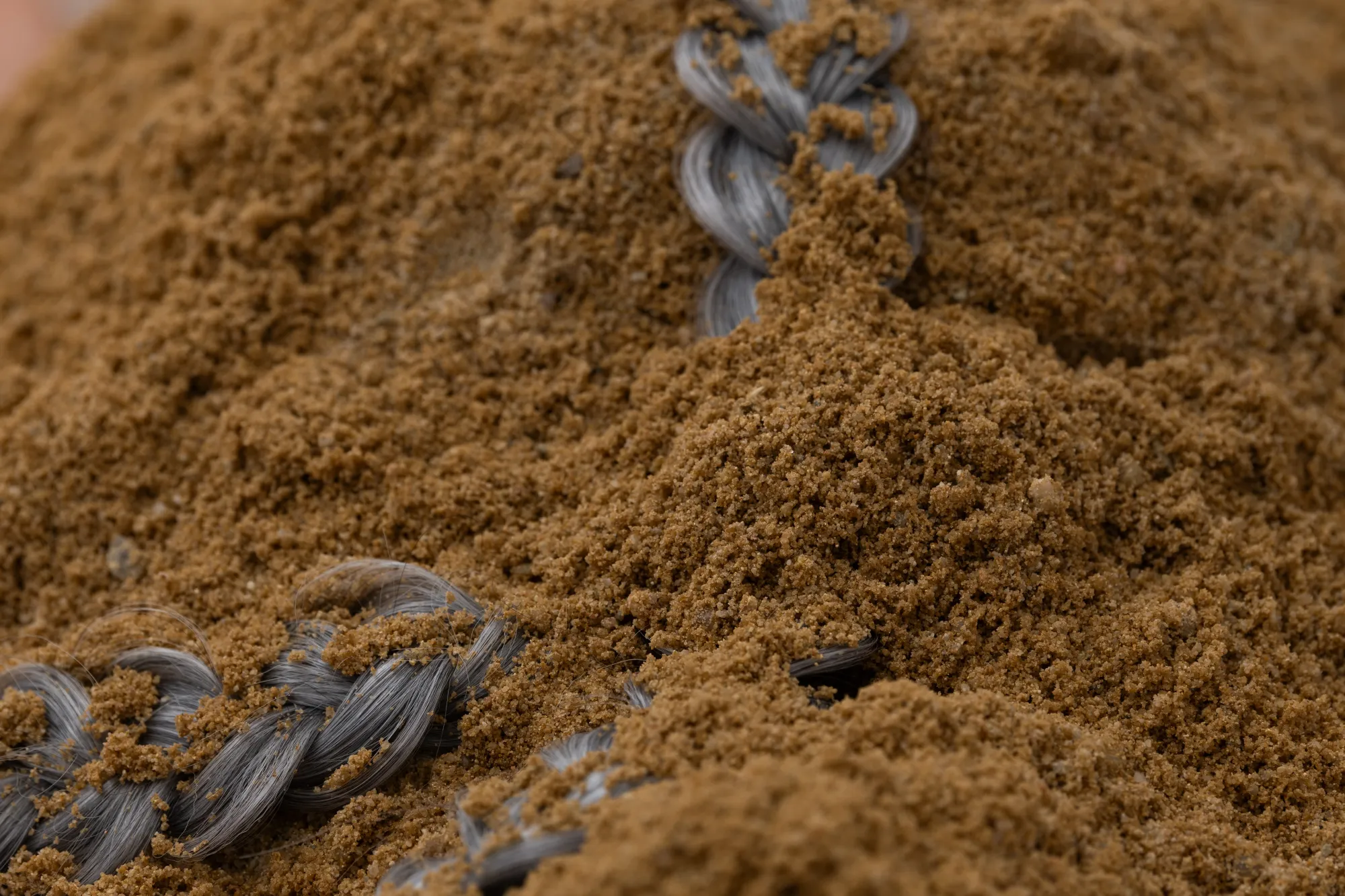 Detail view of Epiphenomenon by Nadine Karl, showing sand partially covering braided gray material embedded in the installation.
