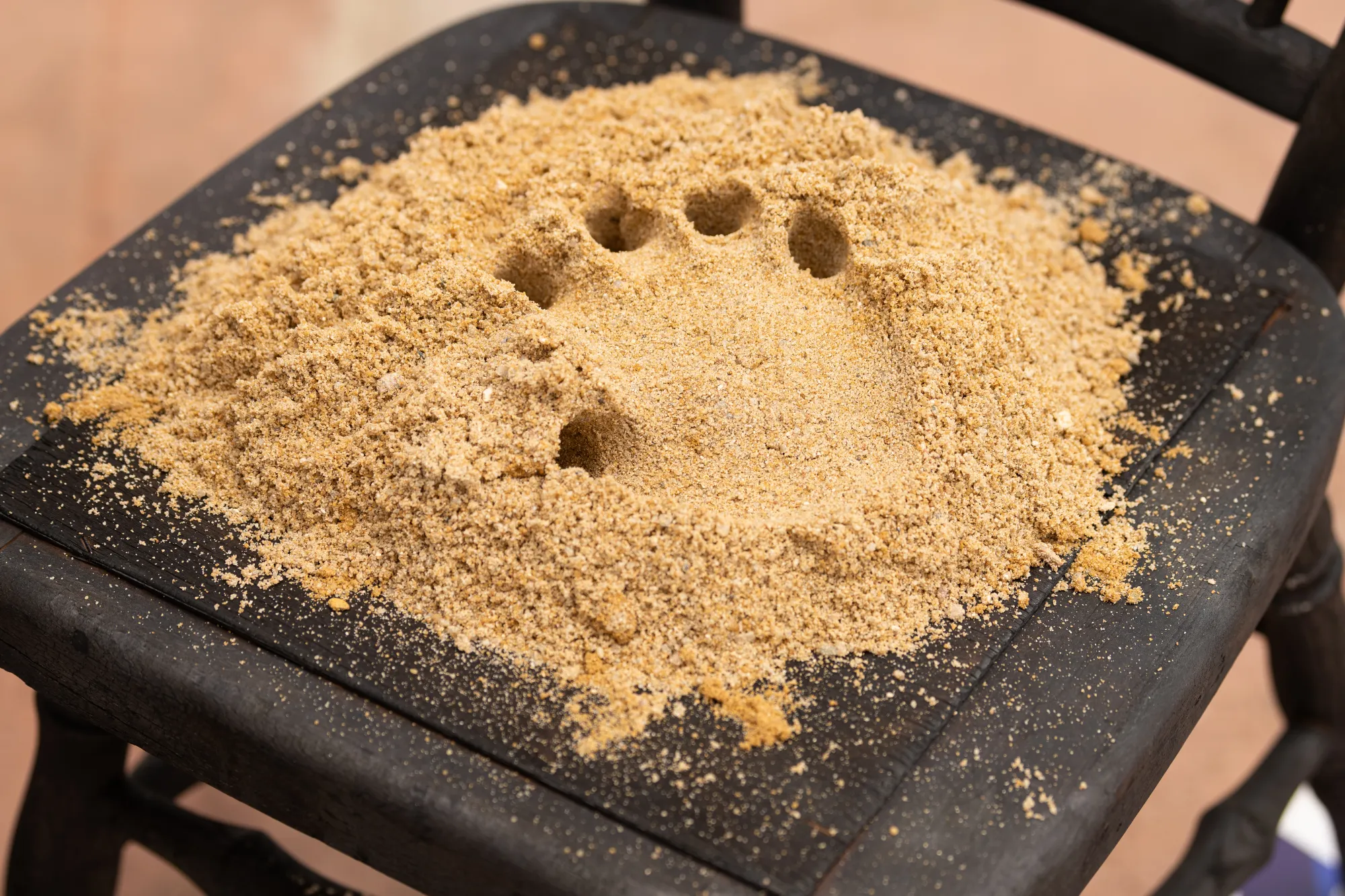 Artist Pages: Detail view of Epiphenomenon by Nadine Karl, showing sand arranged on a wooden chair seat with small circular impressions.