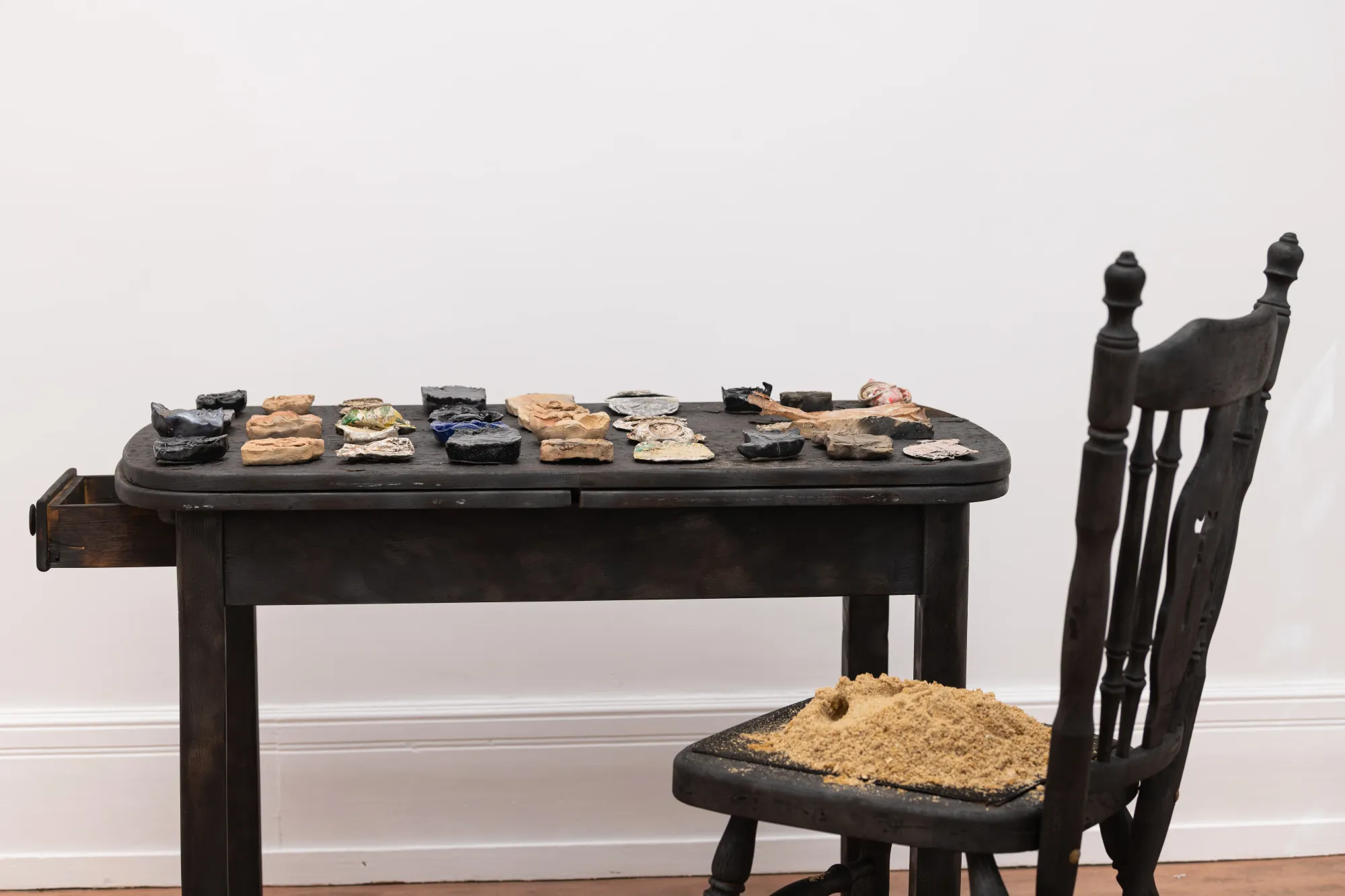 Installation view of Epiphenomenon by Nadine Karl, featuring a dark wooden table with ceramic and sculptural objects and a chair covered in sand.