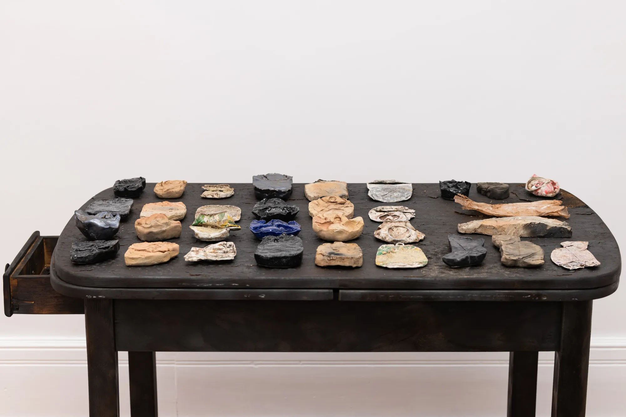 Detail view of Epiphenomenon by Nadine Karl, showing multiple small sculptural and ceramic elements arranged on a dark wooden table.