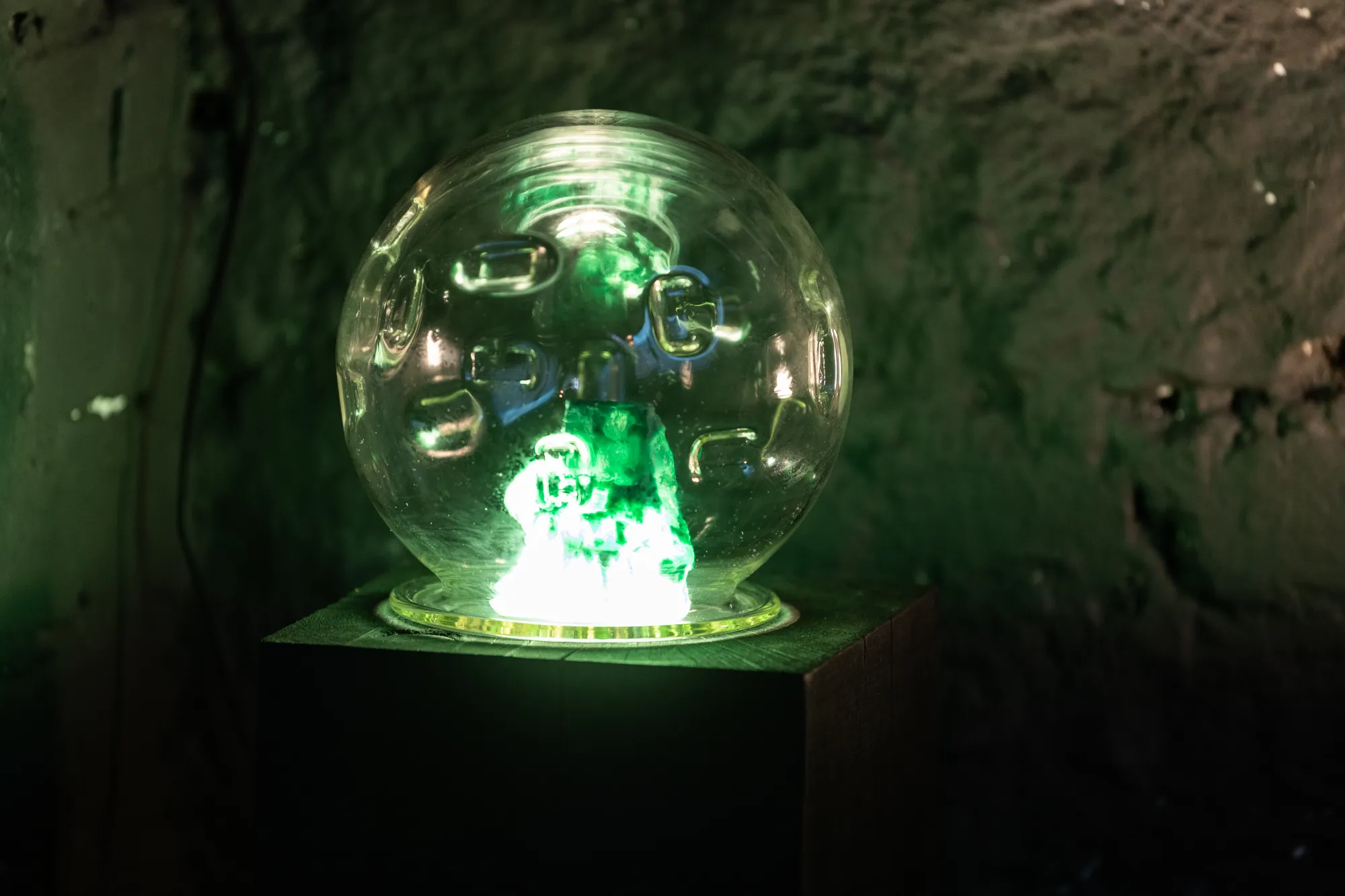Installation detail from Epiphenomenon by Nadine Karl, showing a glass object illuminated with green light placed on a dark pedestal.