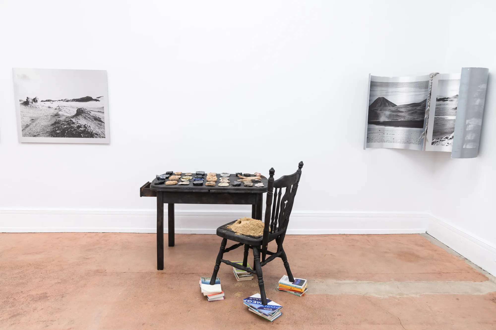 Installation view of Nadine Karl’s work Epiphenomenon, showing a table with sculptural elements, a chair, and photographic works mounted on white gallery walls.